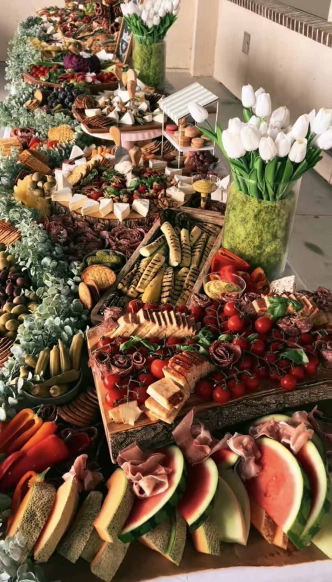 Table with various fruits, vegetables, and flowers, including tulips in a green vase, arranged for display.