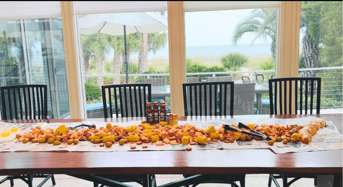Long table with food, large windows showing outdoor patio and greenery, ocean view in the background.
