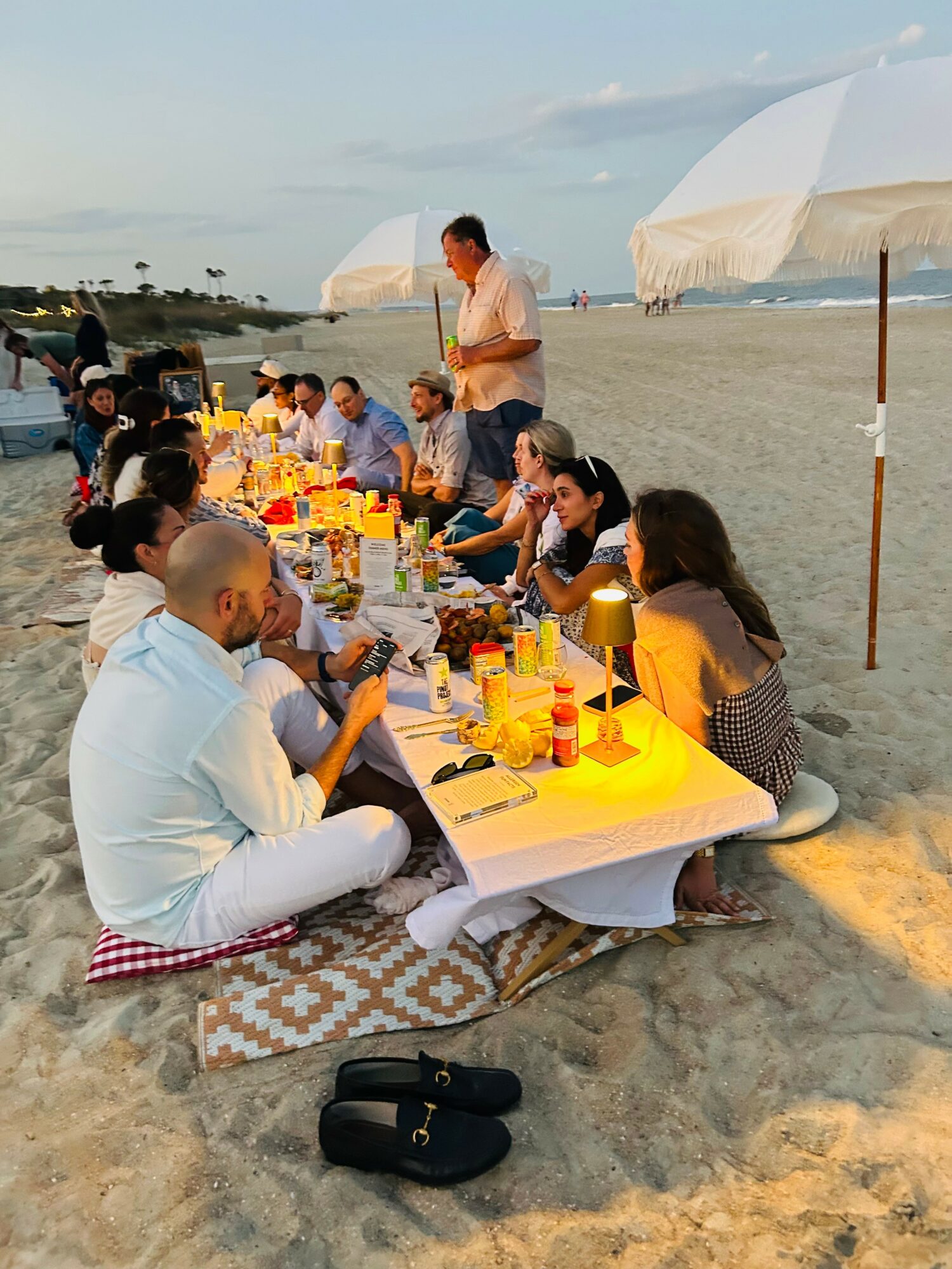 People sit at a long table on the beach with drinks and food, under umbrellas, during sunset. A man stands and speaks.