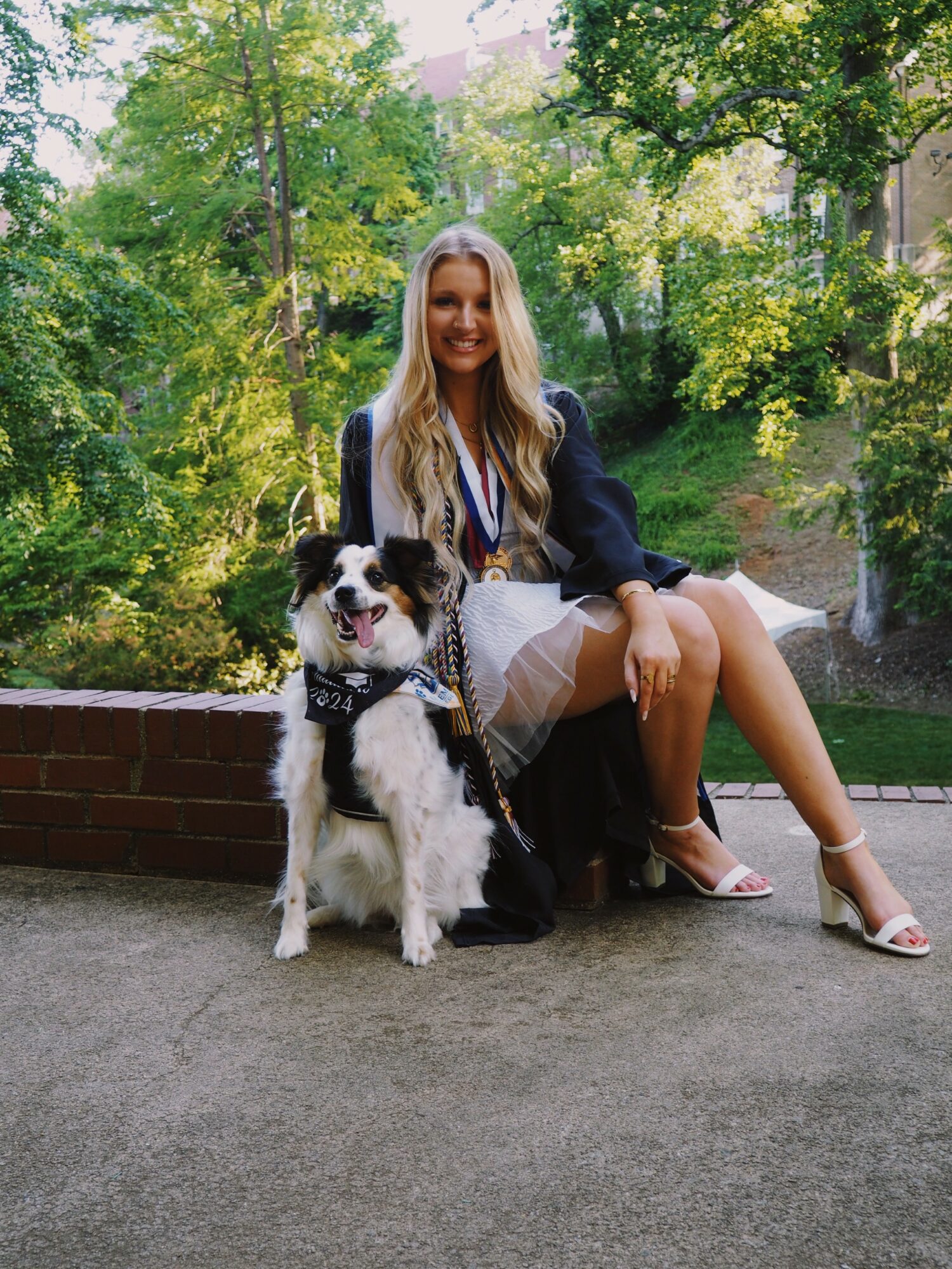 Young woman with long blonde hair sitting outdoors on a brick ledge, smiling, with a black and white dog beside her, surrounded by green trees.