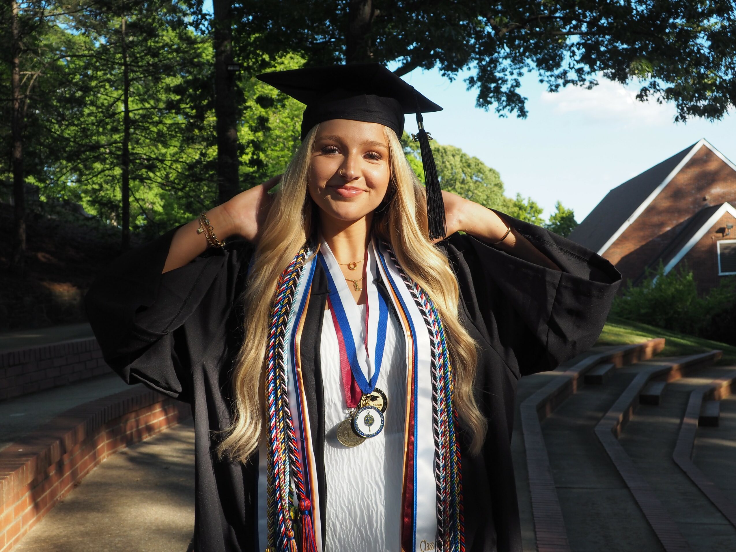 Young woman in graduation cap and gown outdoors, smiling, with trees and a building in background.