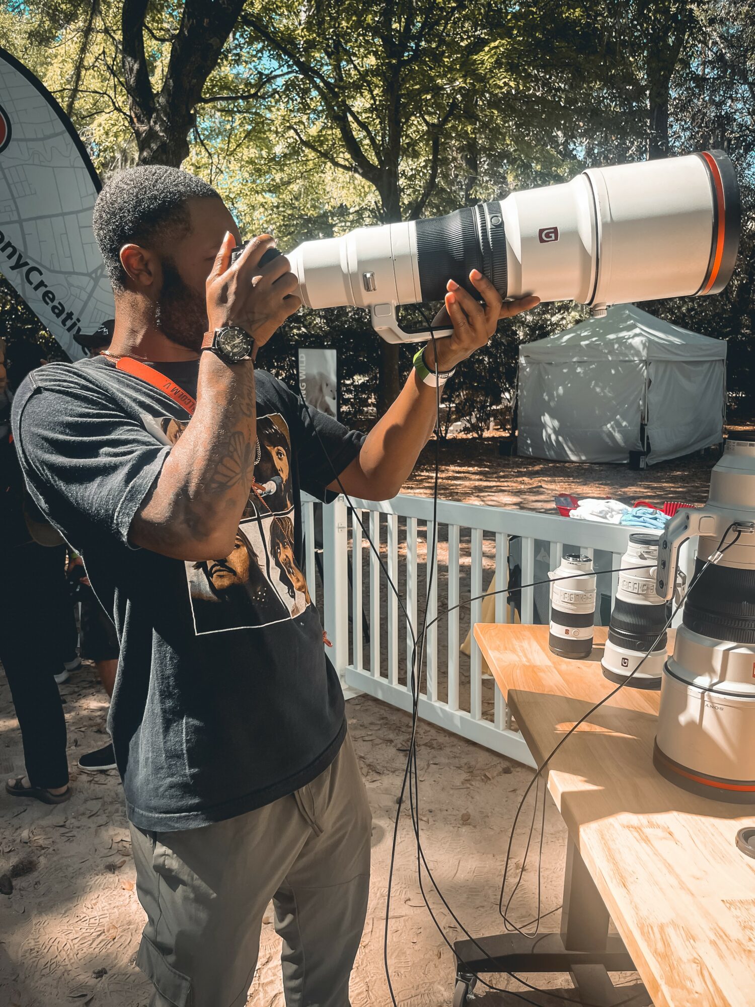 Person holding a large camera lens telescope outdoors, with trees and a table with camera equipment nearby.