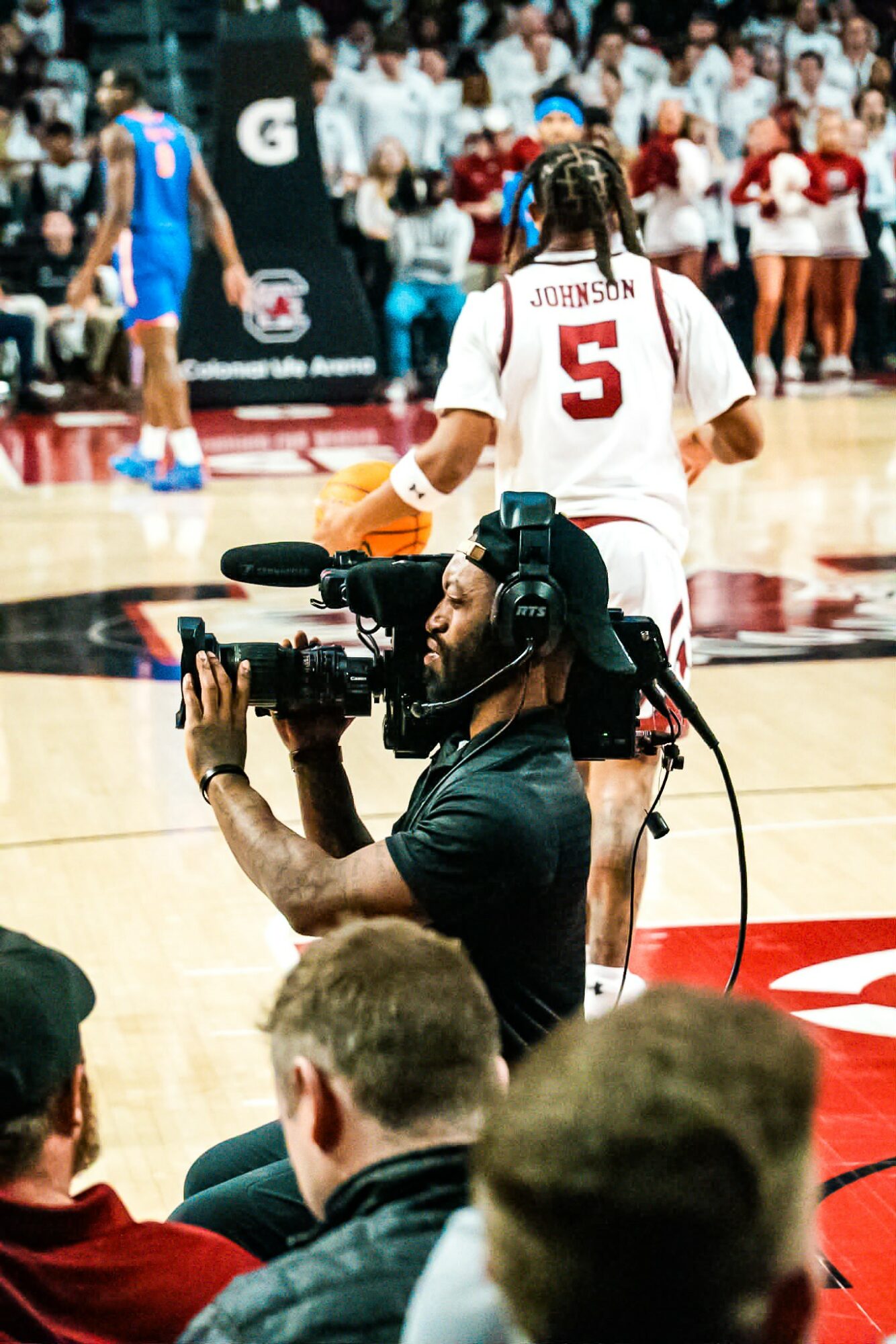 Basketball game scene with player wearing number 5 jersey, camera operator filming, and audience in background.