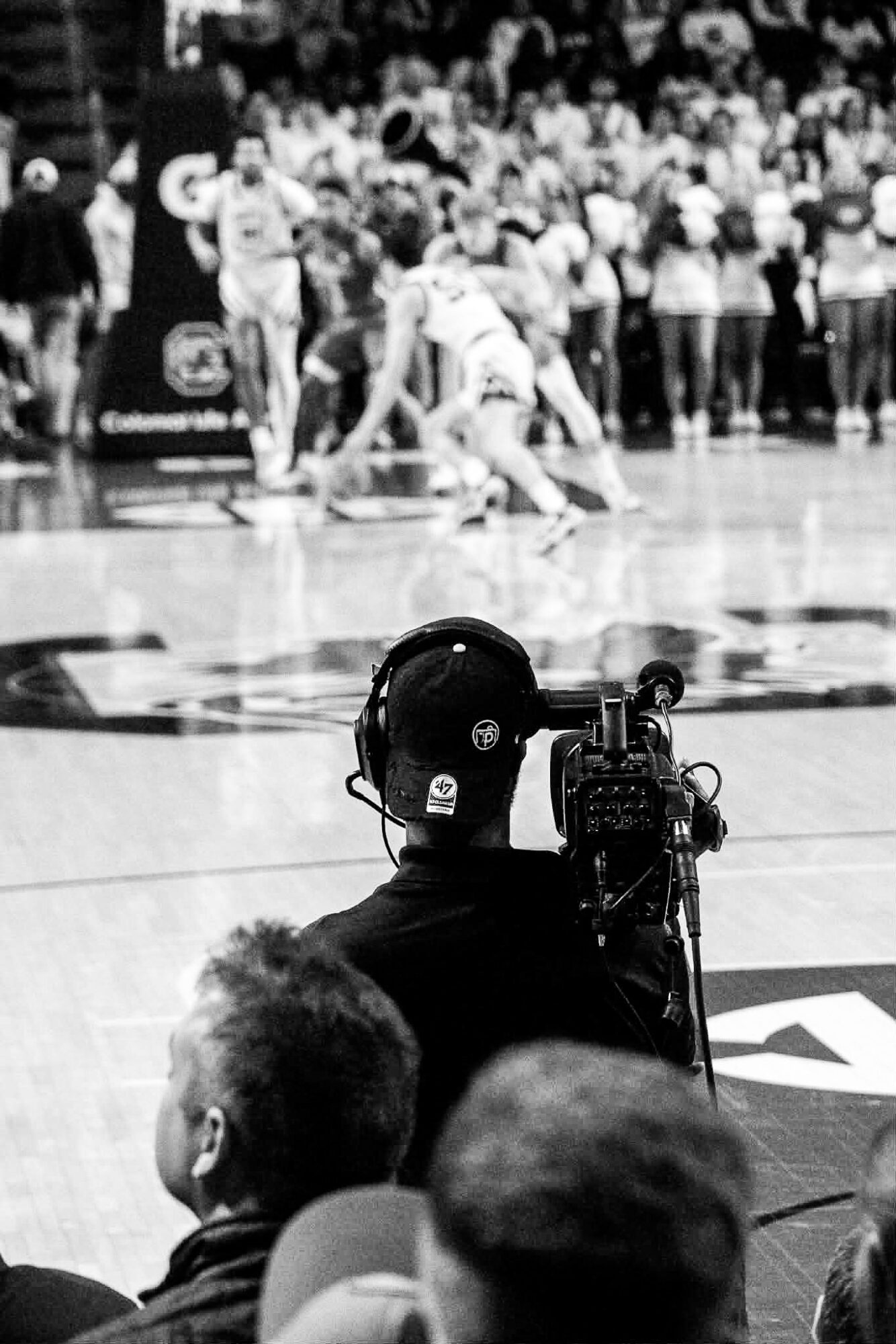 A person with a camera records a basketball game with players on the court and spectators in the background.