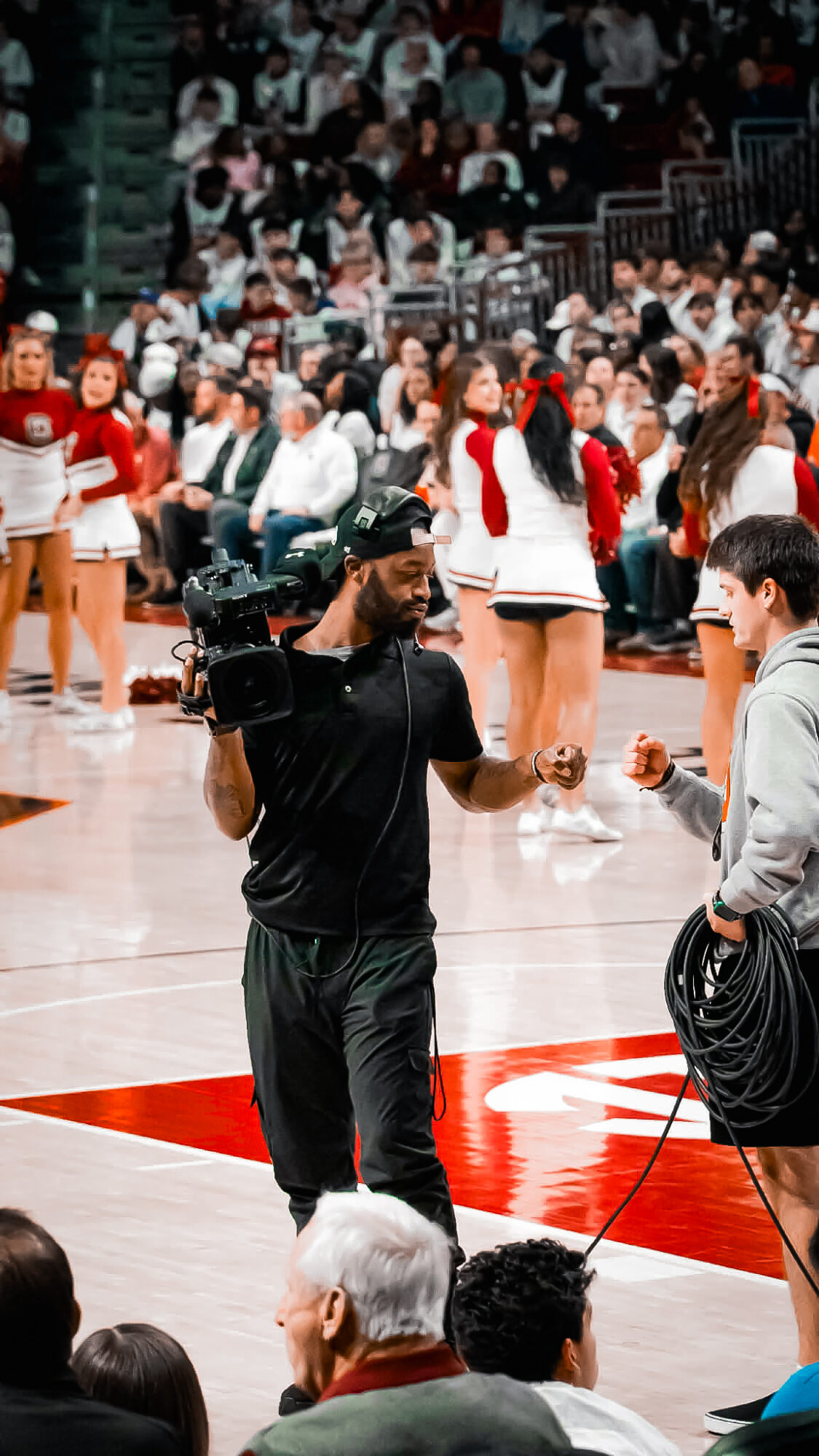 Person in black clothing holding a camera and talking to a young man on a basketball court, with spectators in the background.
