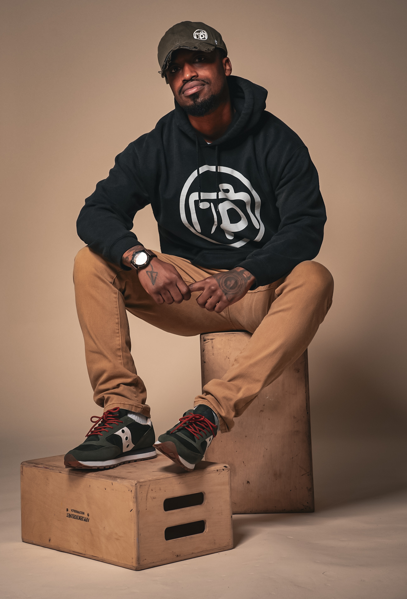 Man sitting on a wooden box, wearing a black hoodie, tan pants, sneakers, and a cap, smiling at the camera.