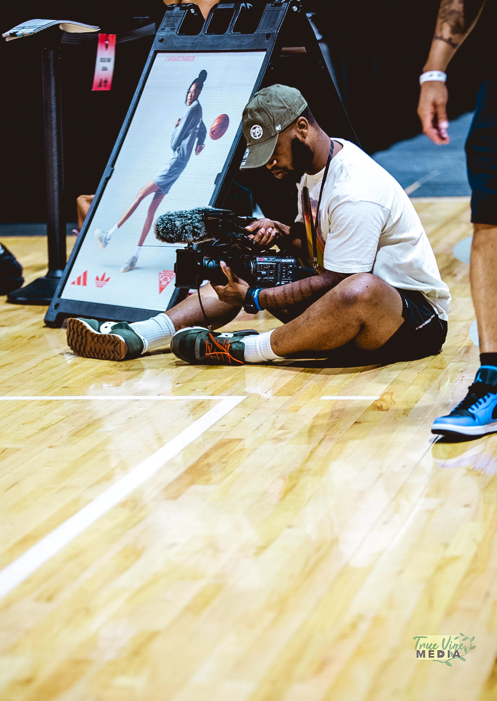 Person sitting on the floor using a camera, with a large monitor displaying a digital illustration of a character, on a wooden floor.
