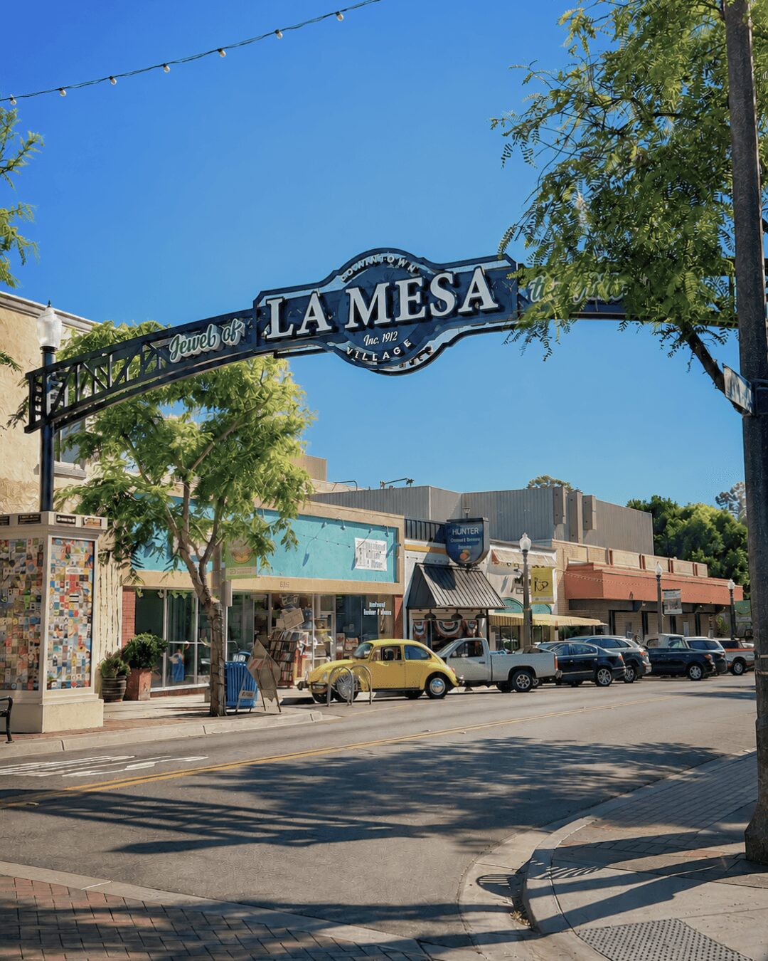 Street view with storefronts, parked cars, trees, and a sign reading 'LA MESA' against a clear blue sky.