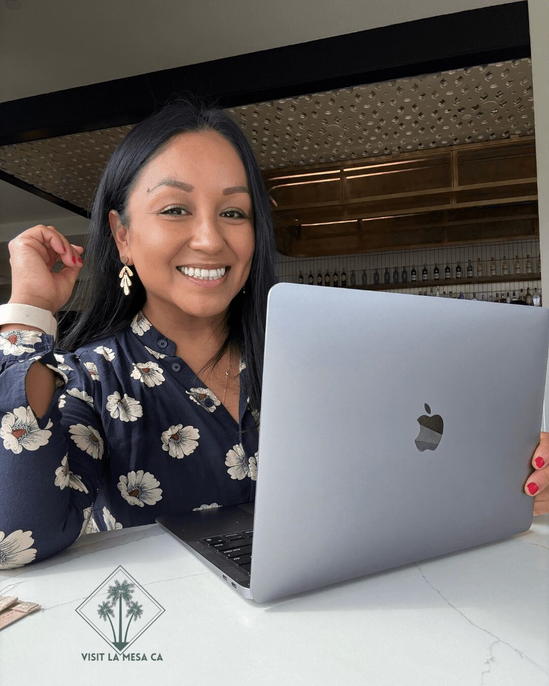 Woman smiling at laptop, wearing floral blouse, sitting at table indoors.