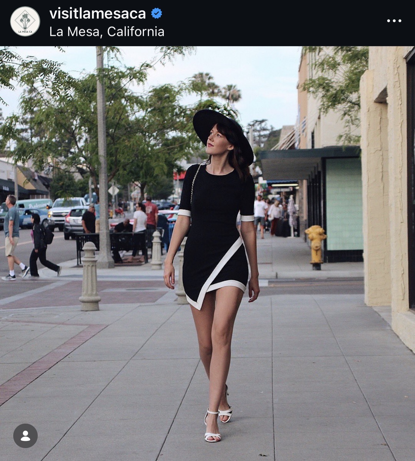 Woman walking on sidewalk wearing black dress, large hat, and high heels, with trees and buildings in background.