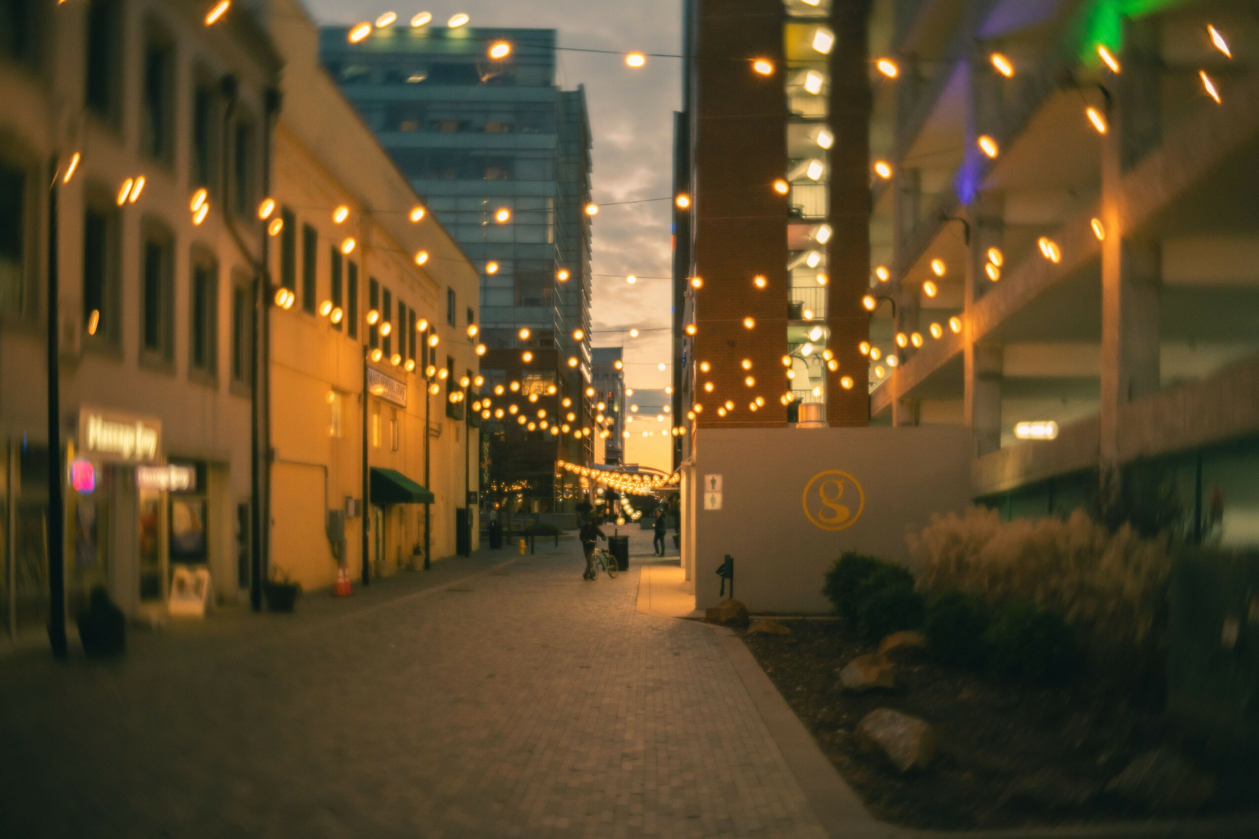 Street decorated with string lights, buildings on both sides, and a few people walking, during evening or night.