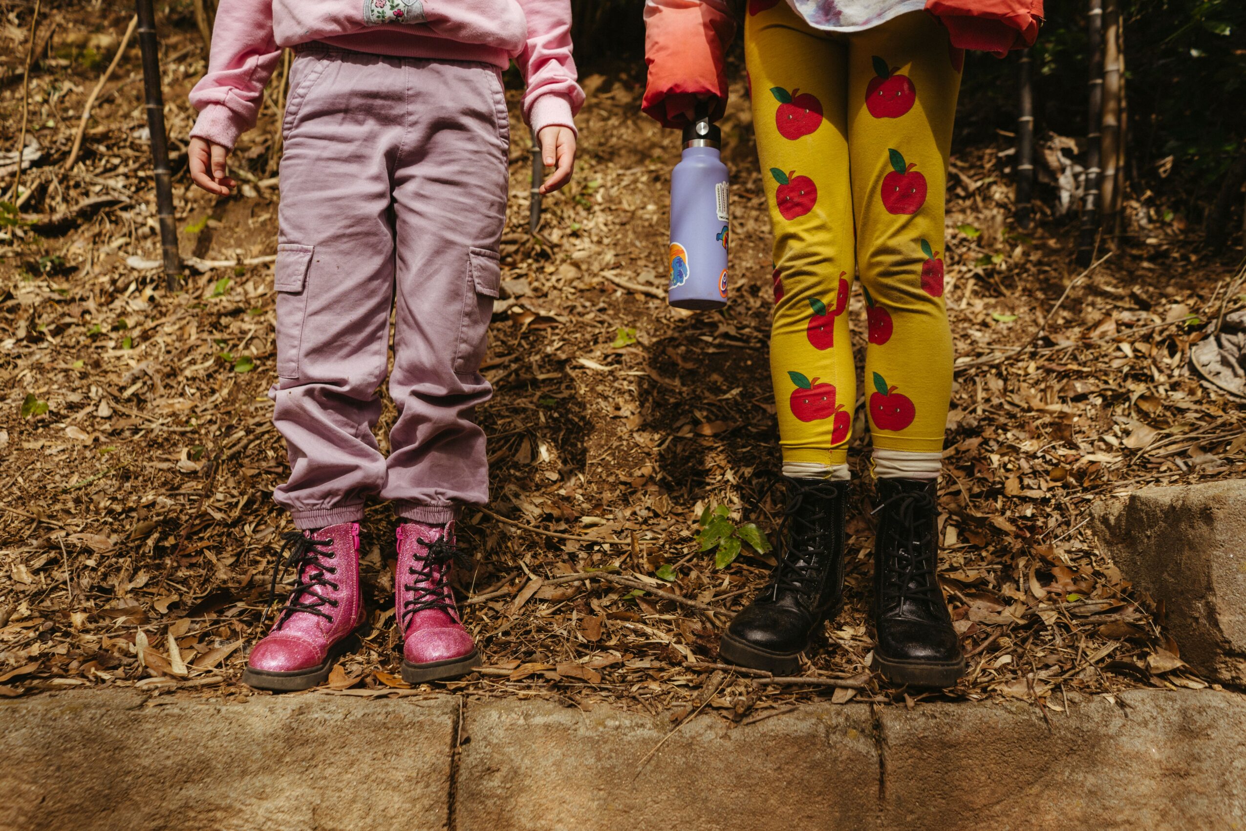 Two children stand outdoors on a dirt path, wearing colorful pants and boots, with trees in the background.