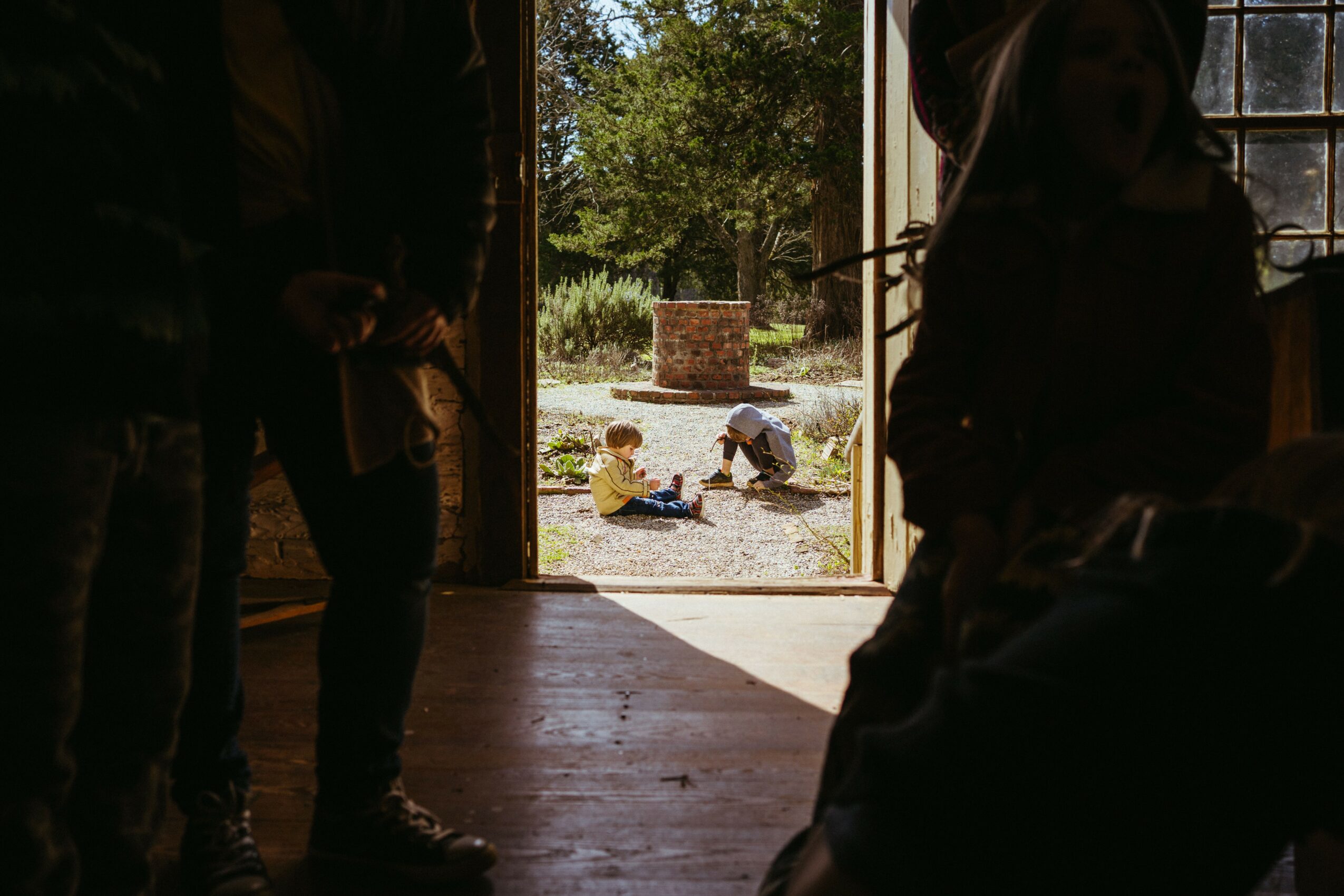 People inside a building looking out at children playing outdoors in sunlight, with trees and a brick structure visible.