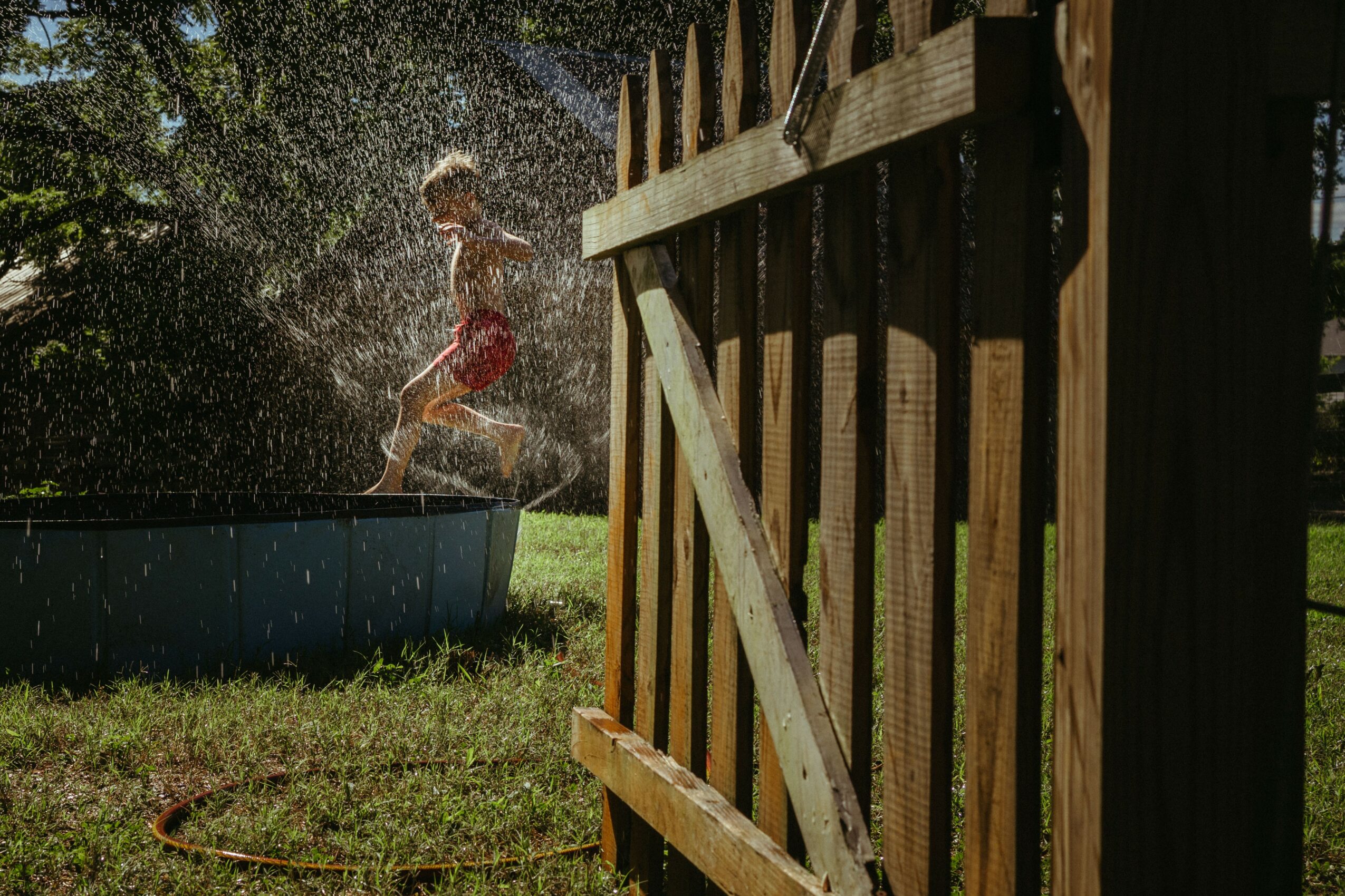 Child playing in a splash pool outdoors, water splashing, wooden fence partially visible, grassy area, sunlight, and trees in background.