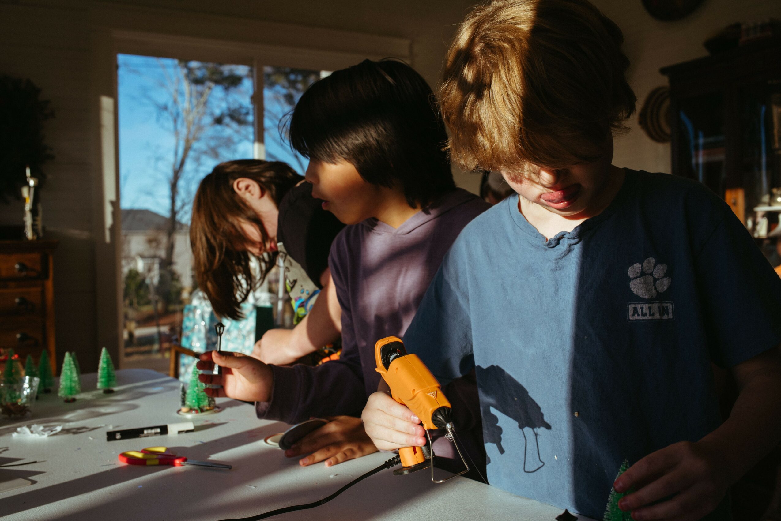 Three children playing with toys and tools at a table indoors, sunlight streaming through a window behind them.