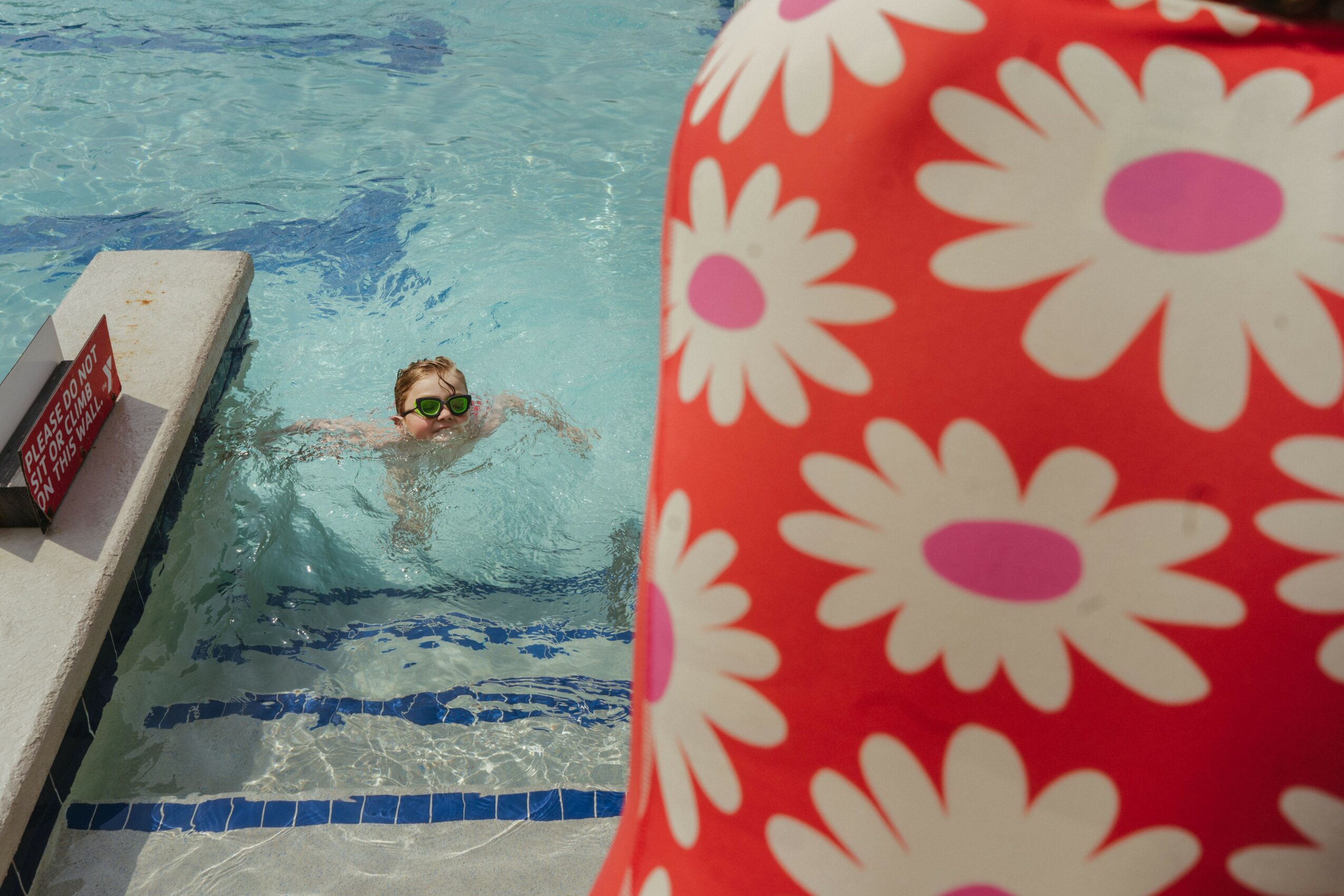 Child swimming in a pool with goggles, partially obscured by a person wearing a red and white floral swimsuit, pool edge visible.