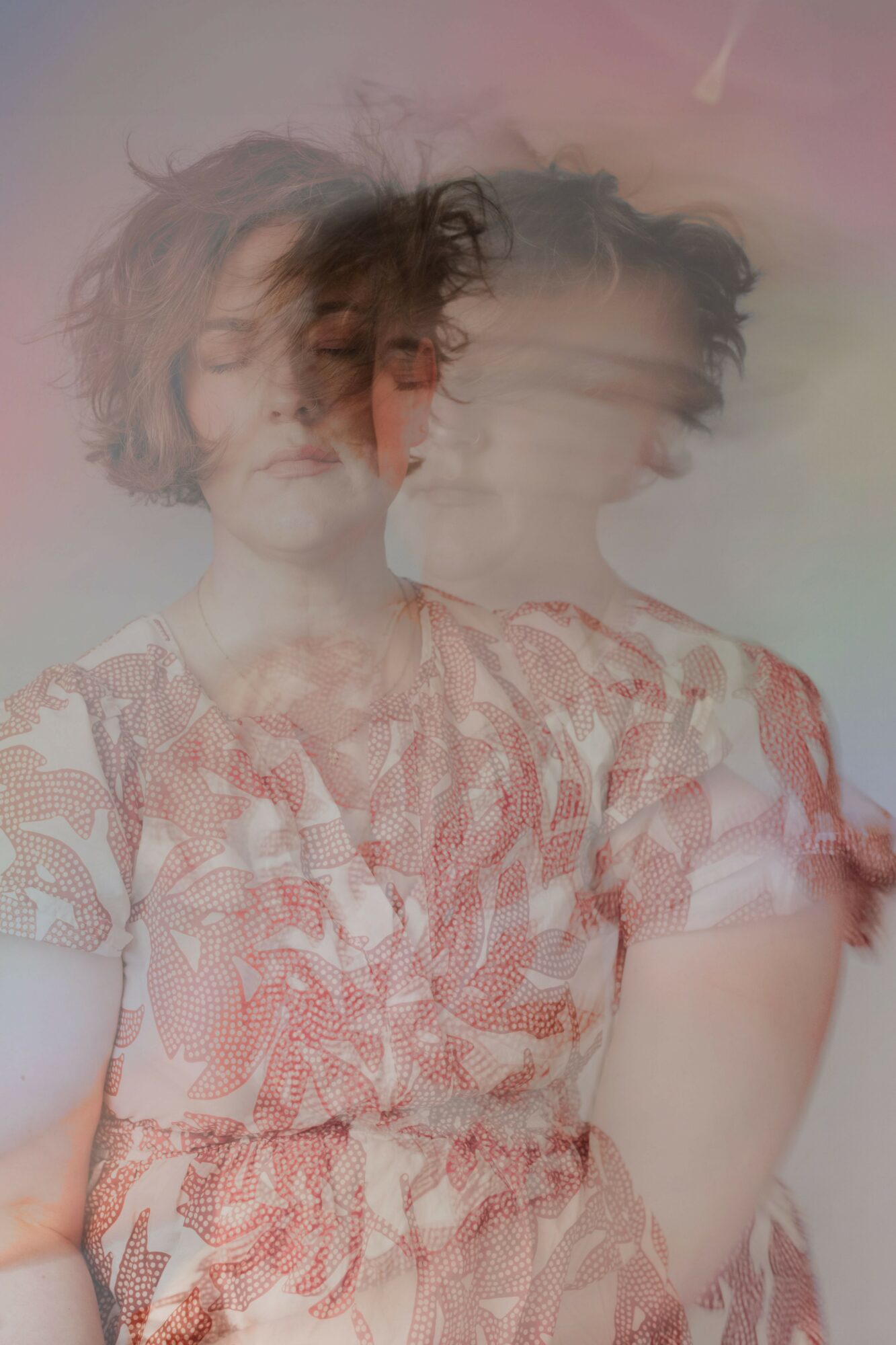 Two women with short, curly hair wearing patterned dresses, overlapping and blurred, against a soft pastel background.