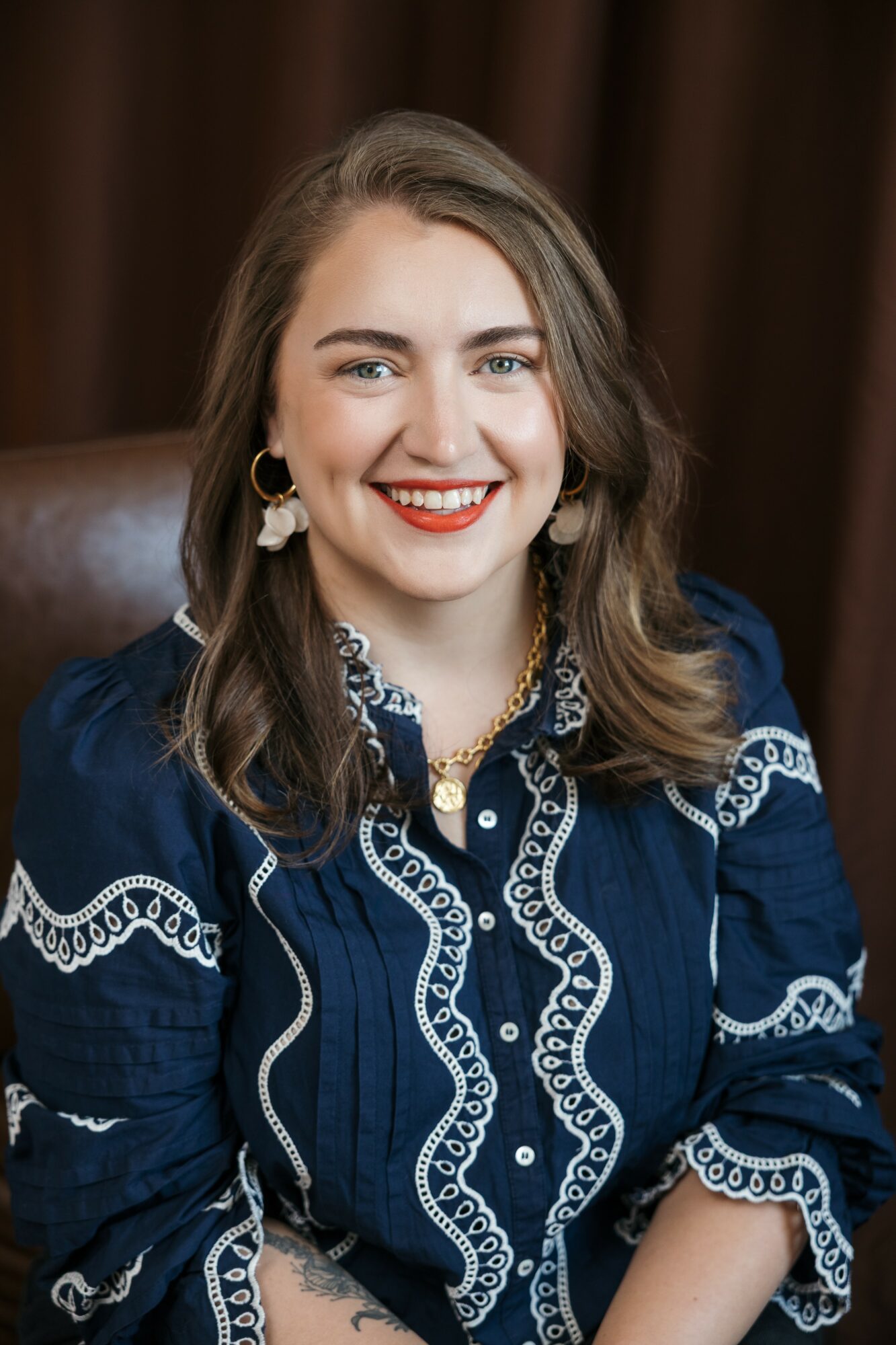 Young woman with long wavy hair smiling, wearing a navy blue embroidered blouse, earrings, and a necklace, sitting indoors.