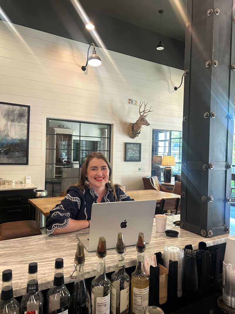 Woman sitting at a bar counter with a laptop, smiling, in a modern cafe with decorative wall art and large windows.