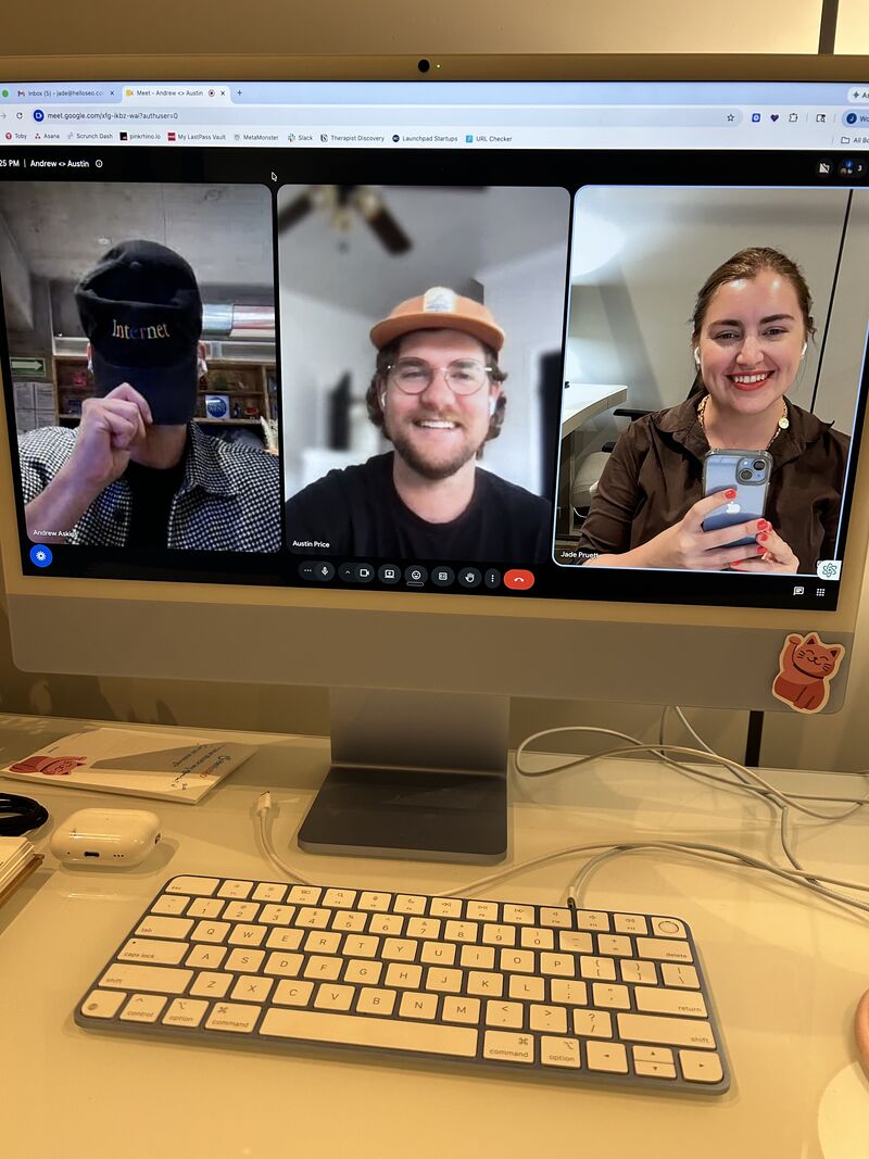 Computer screen showing three people in a video call, smiling and holding phones, with a keyboard and desk items below.