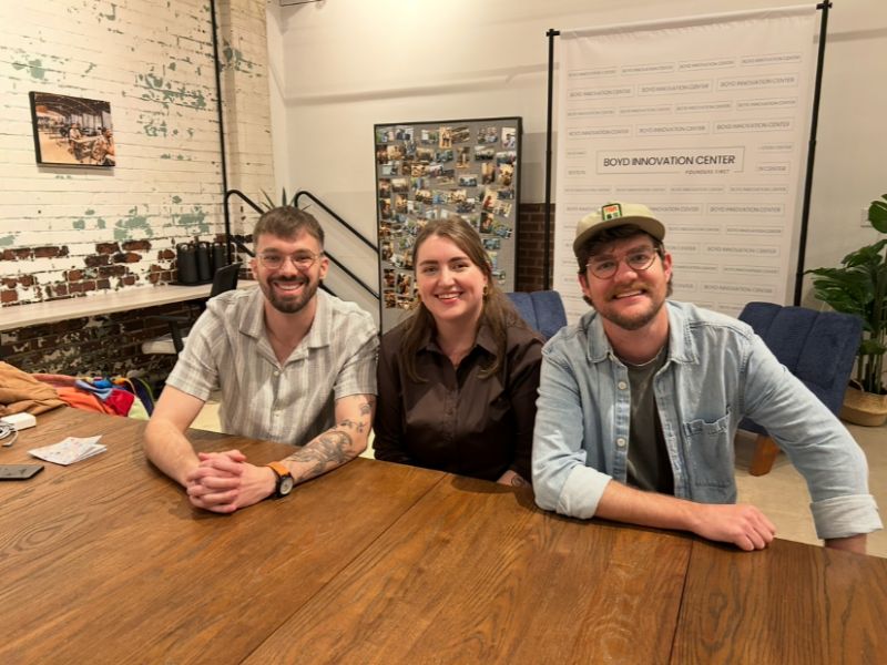 Three people sitting at a wooden table in a room with brick and white walls, smiling at the camera.