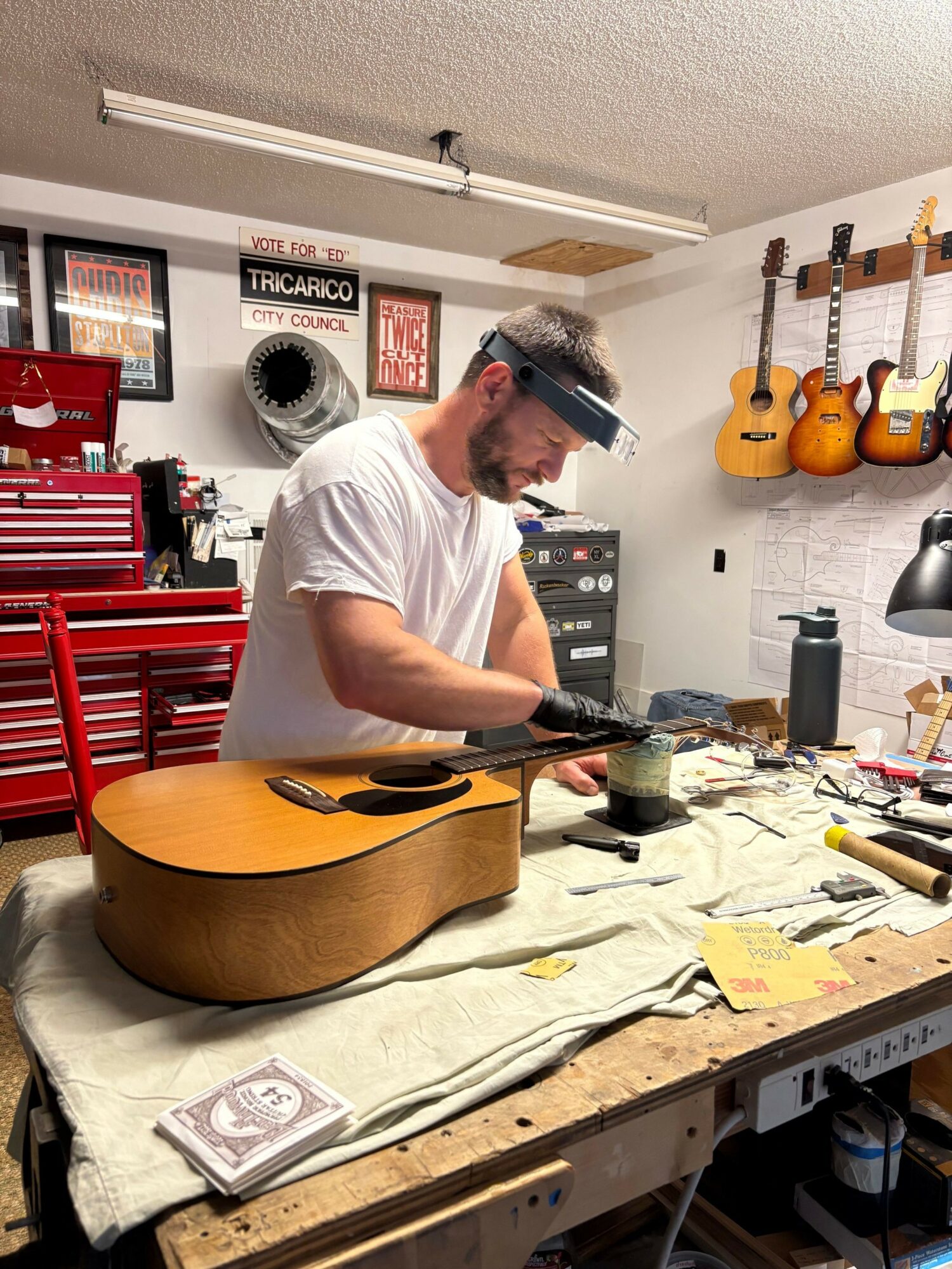 Man with a beard and glasses wearing a white shirt and headphones, working on a guitar in a workshop.