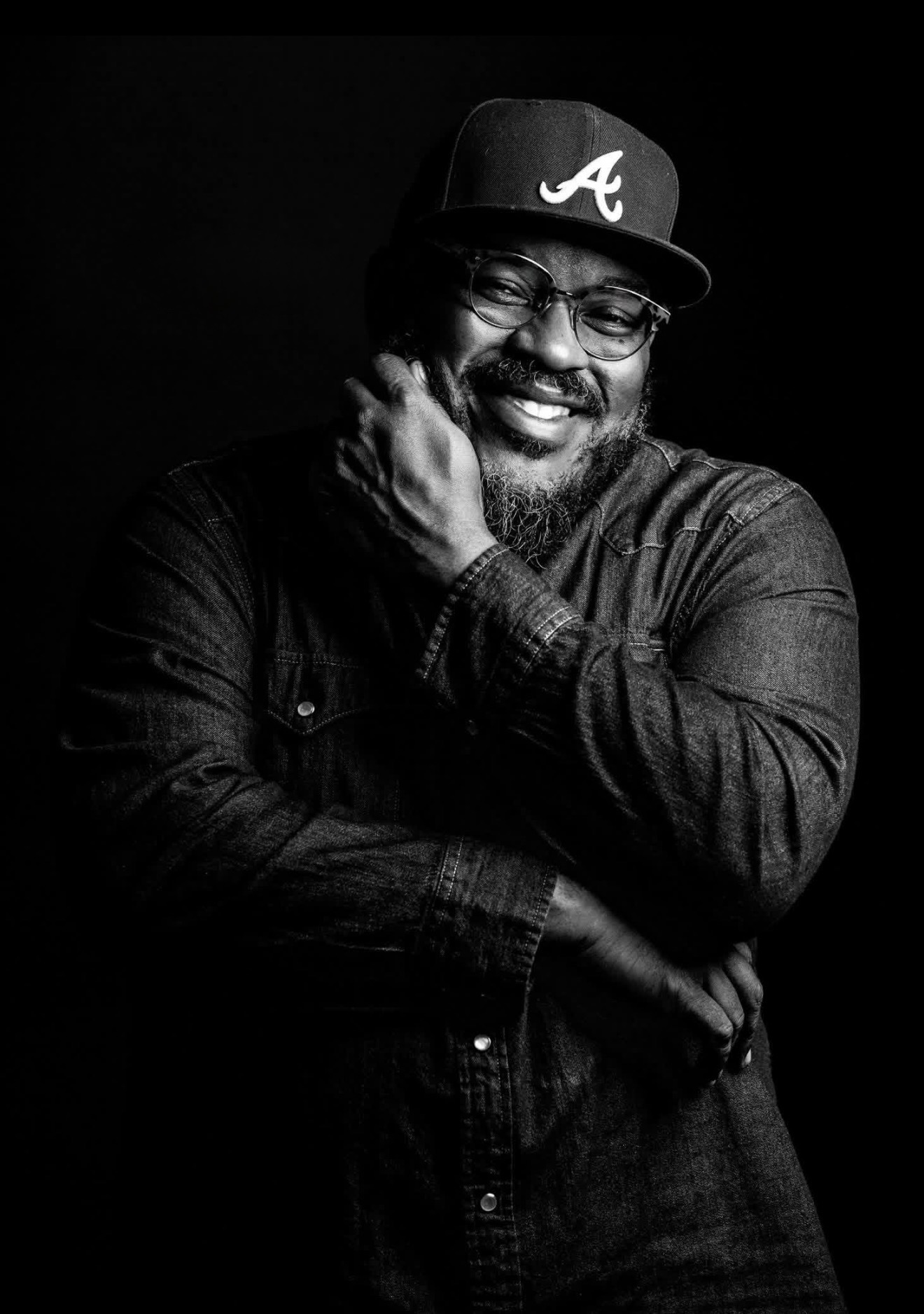 Smiling man wearing glasses and a baseball cap with an 'A' logo, touching his face with one hand, in black and white.
