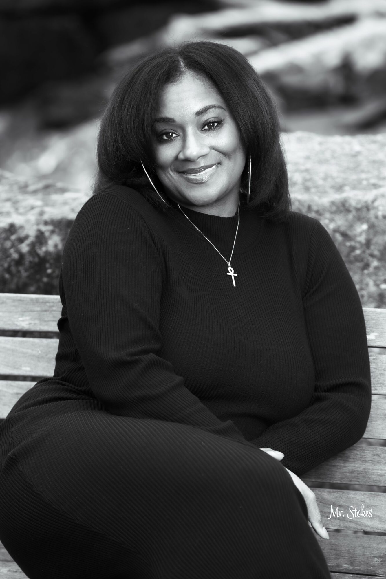 Woman with shoulder-length hair smiling, sitting outdoors on a bench, wearing a dark top and cross necklace.