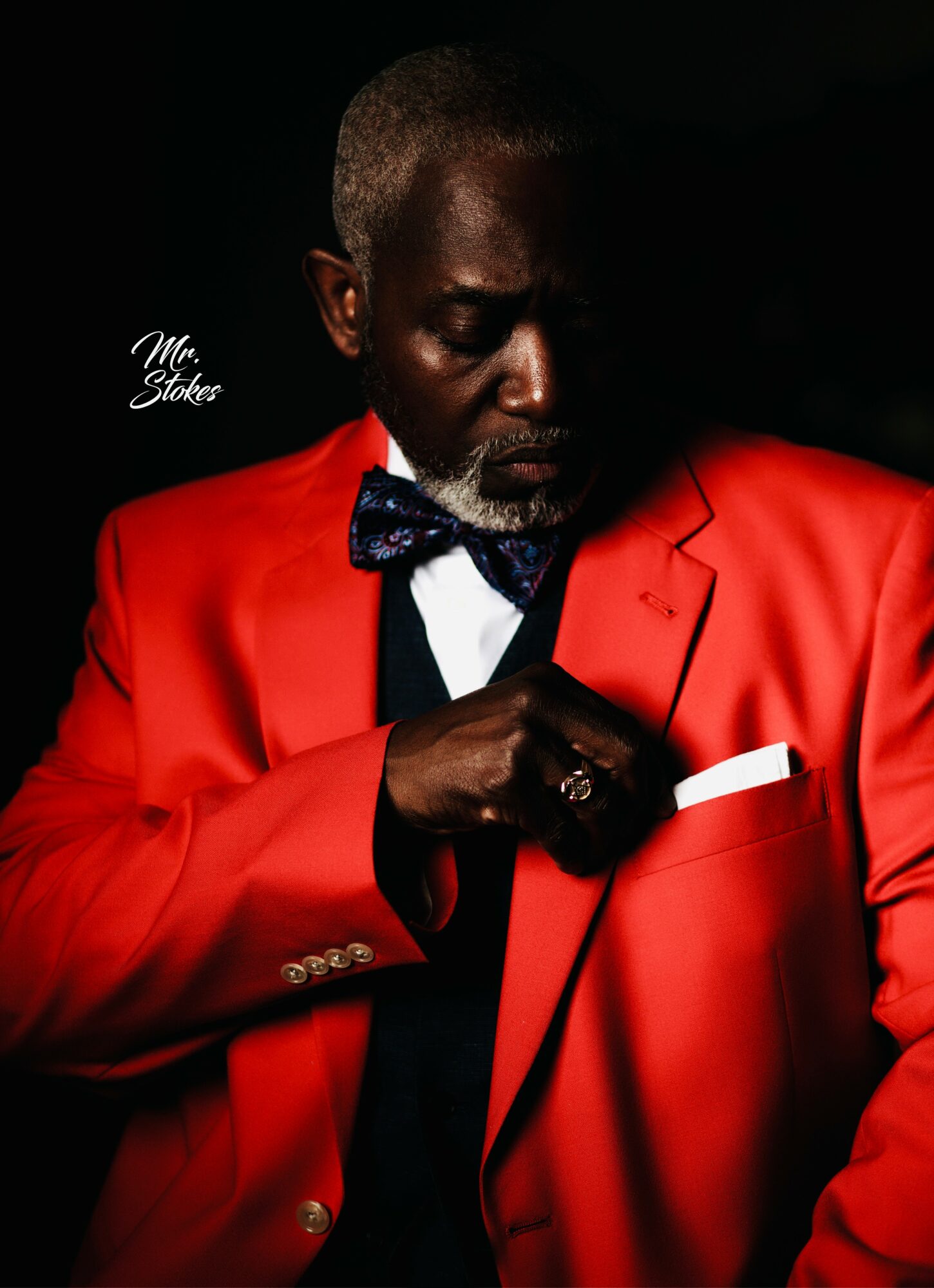 Man in a red suit with a bow tie, adjusting a white pocket square, dark background, serious expression.