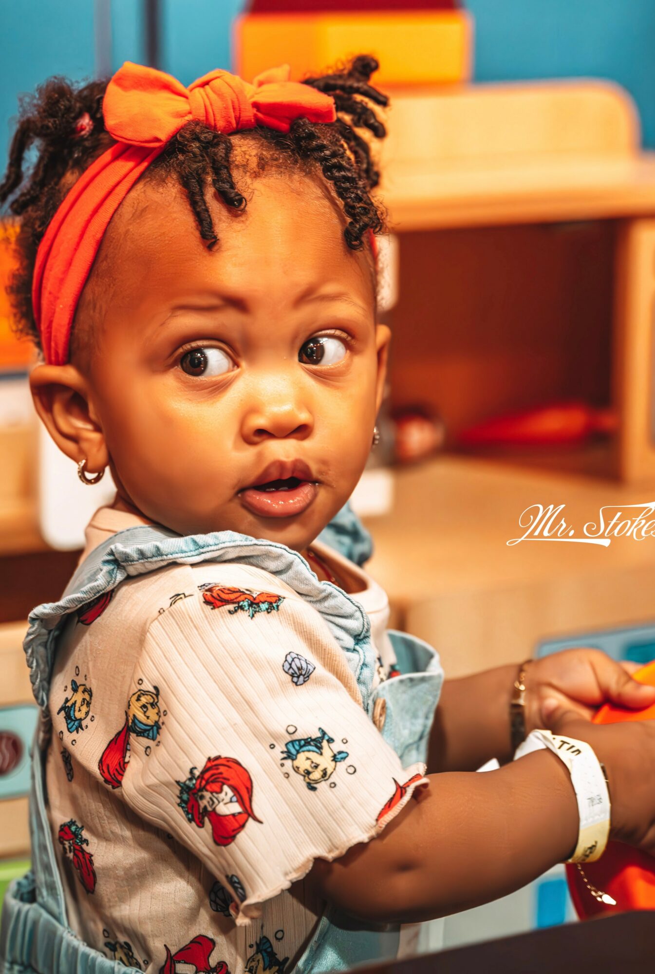 Young girl with a red headband and curly hair, looking to the side, in a colorful indoor setting.