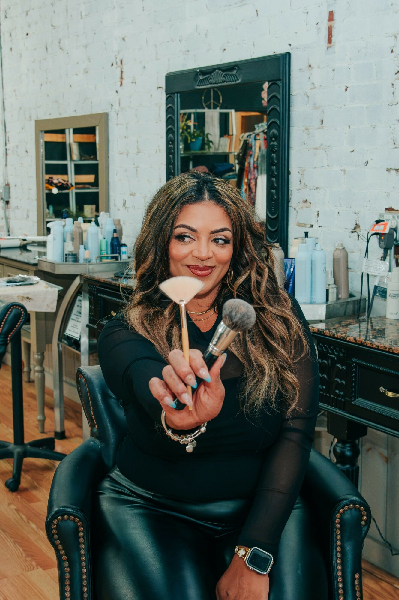 Woman with long hair holding makeup brushes in a salon, smiling, with mirrors and beauty products in background.