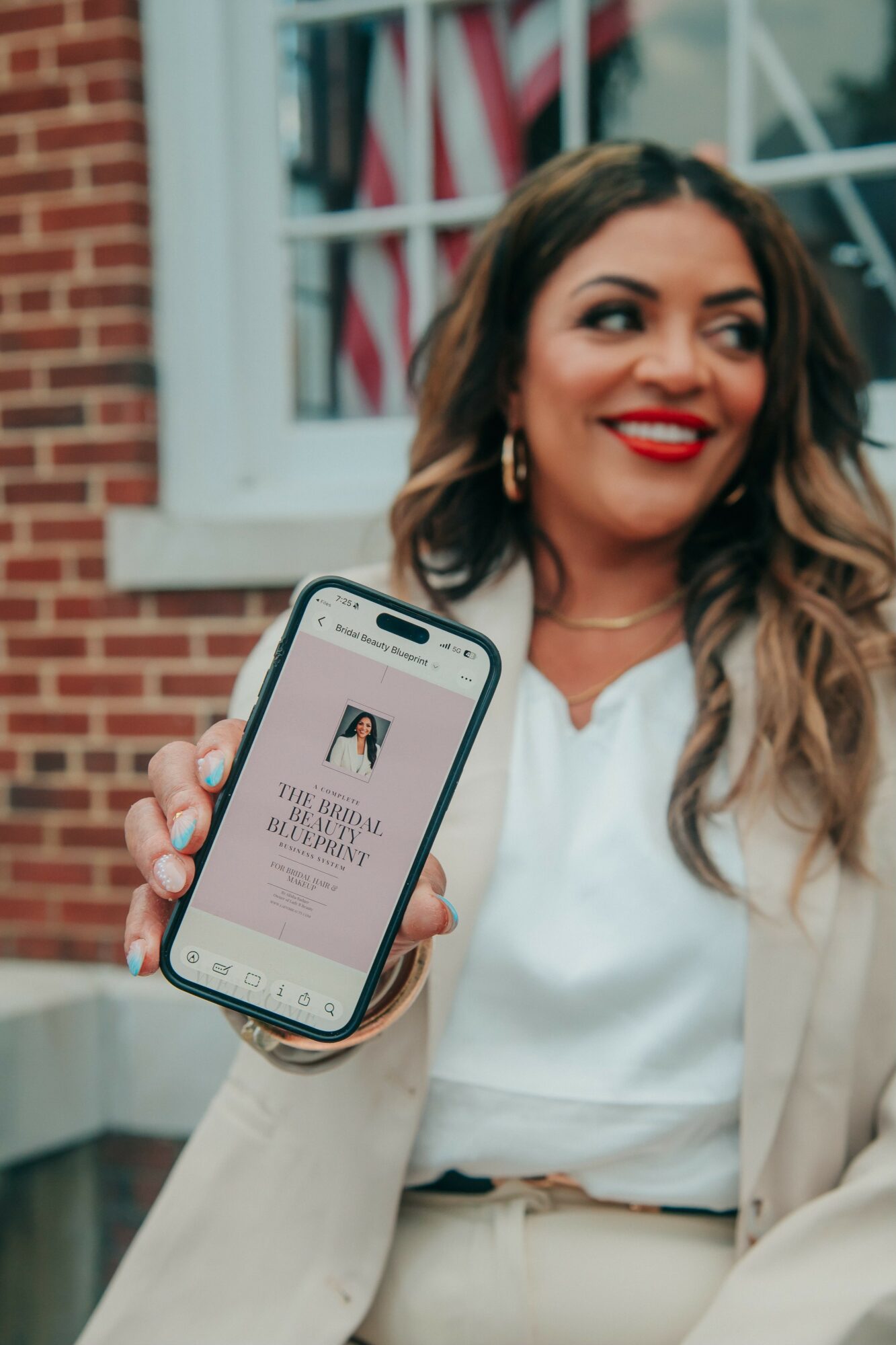 Woman with long hair and red lipstick holding a smartphone showing a message, standing outside a brick building with a window and American flag.