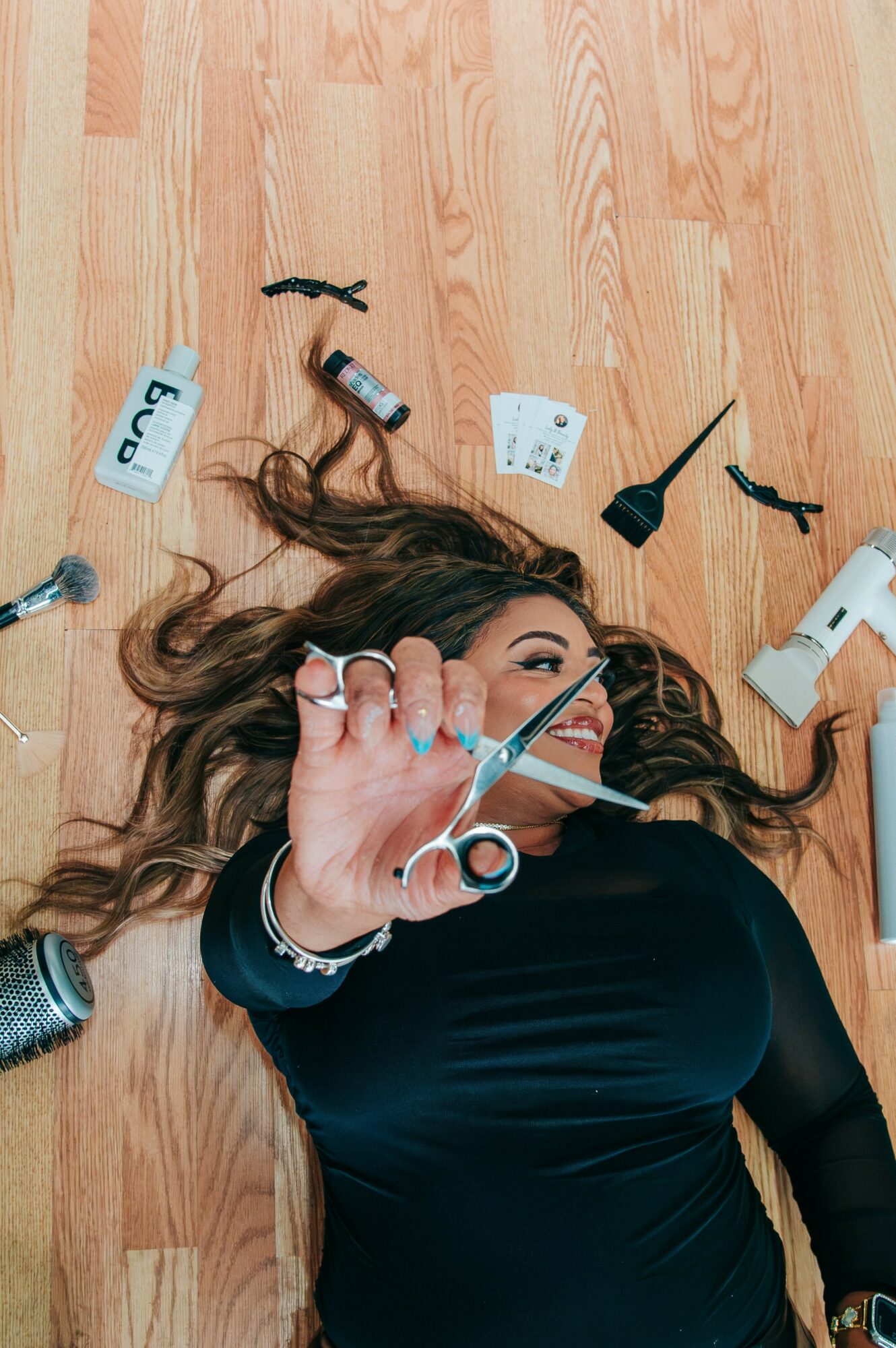 Woman lying on wooden floor with hair spread, surrounded by hairdressing tools and accessories, holding scissors towards camera.