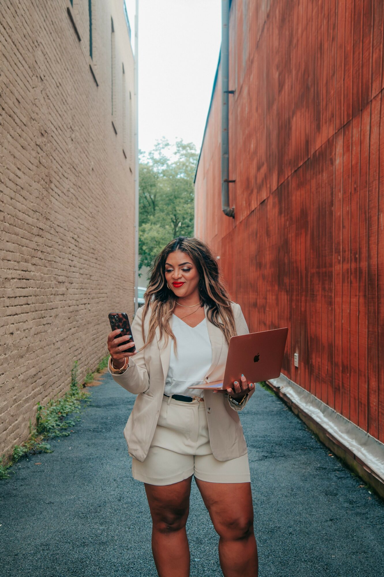 Woman holding a smartphone and a laptop in a narrow alleyway between two buildings, one beige and one red.