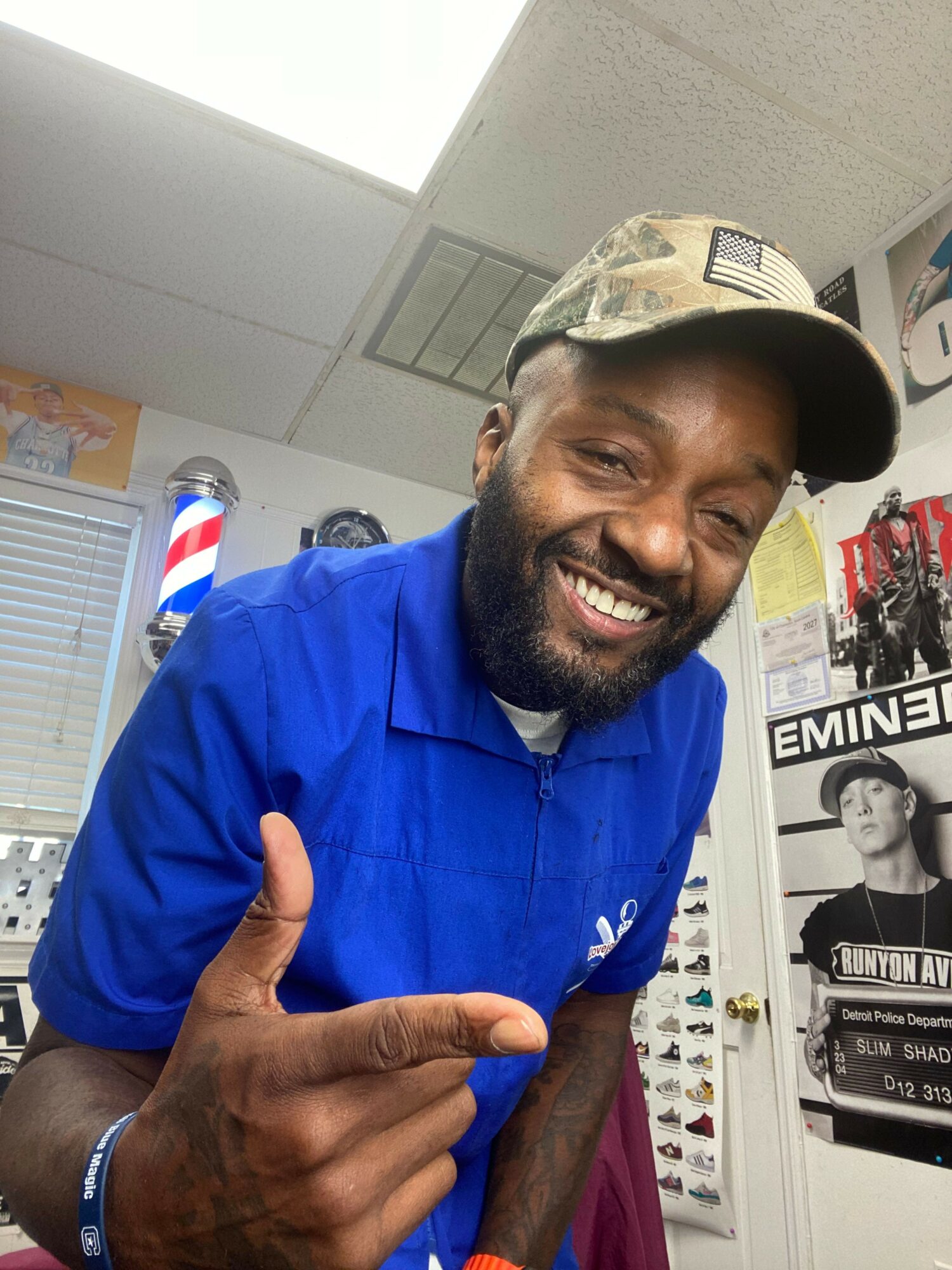 Man smiling, wearing a cap and blue shirt, making a hand gesture, in a room with posters and shelves.