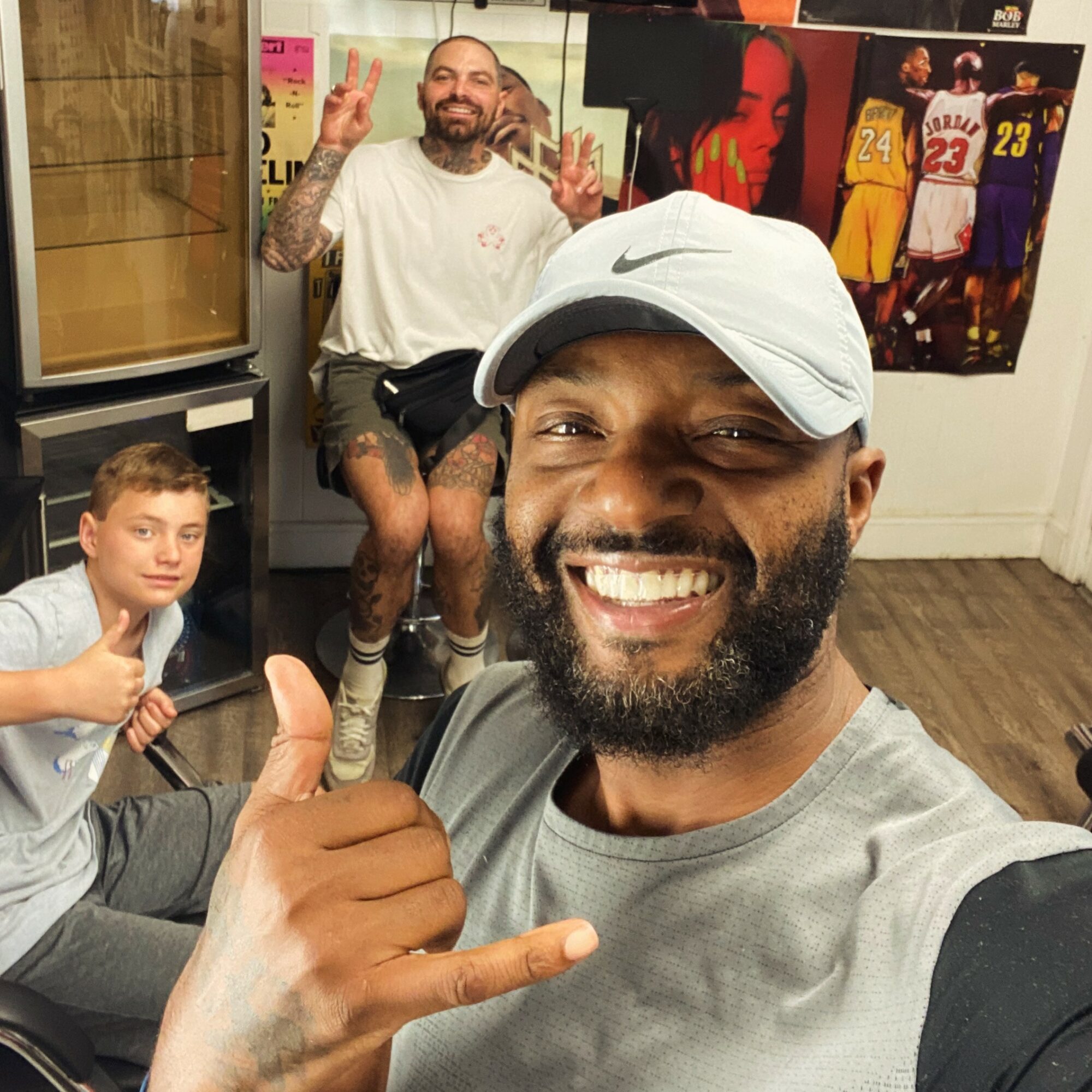 Two men and a boy in a room with sports posters, smiling and making gestures, one in a white cap taking a selfie.