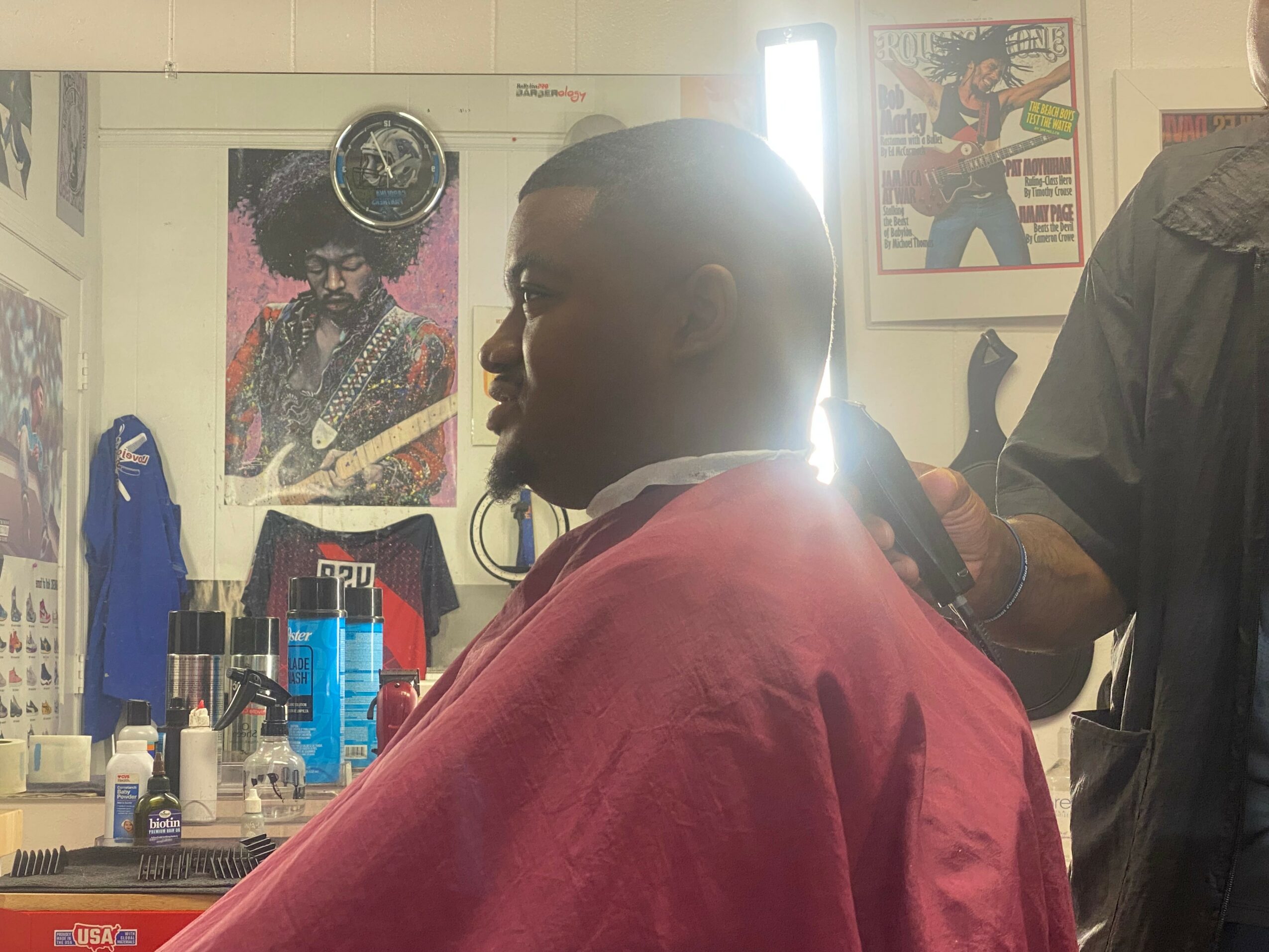 Person sitting in a barber shop chair with a red cape, facing left, with posters and products in the background.