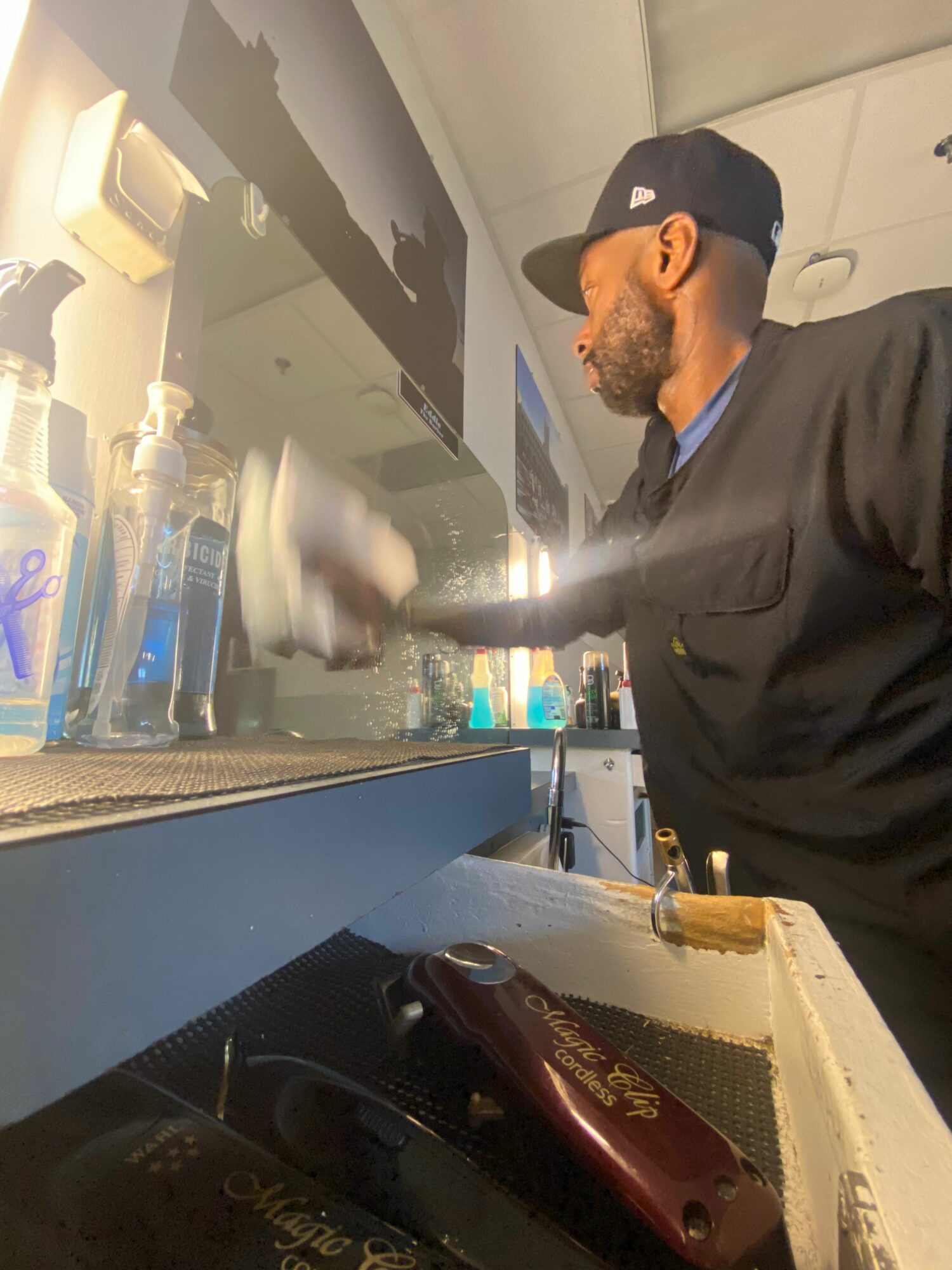 Man with beard and cap cleaning a glass display case in a store, with various bottles inside, on a blue counter.