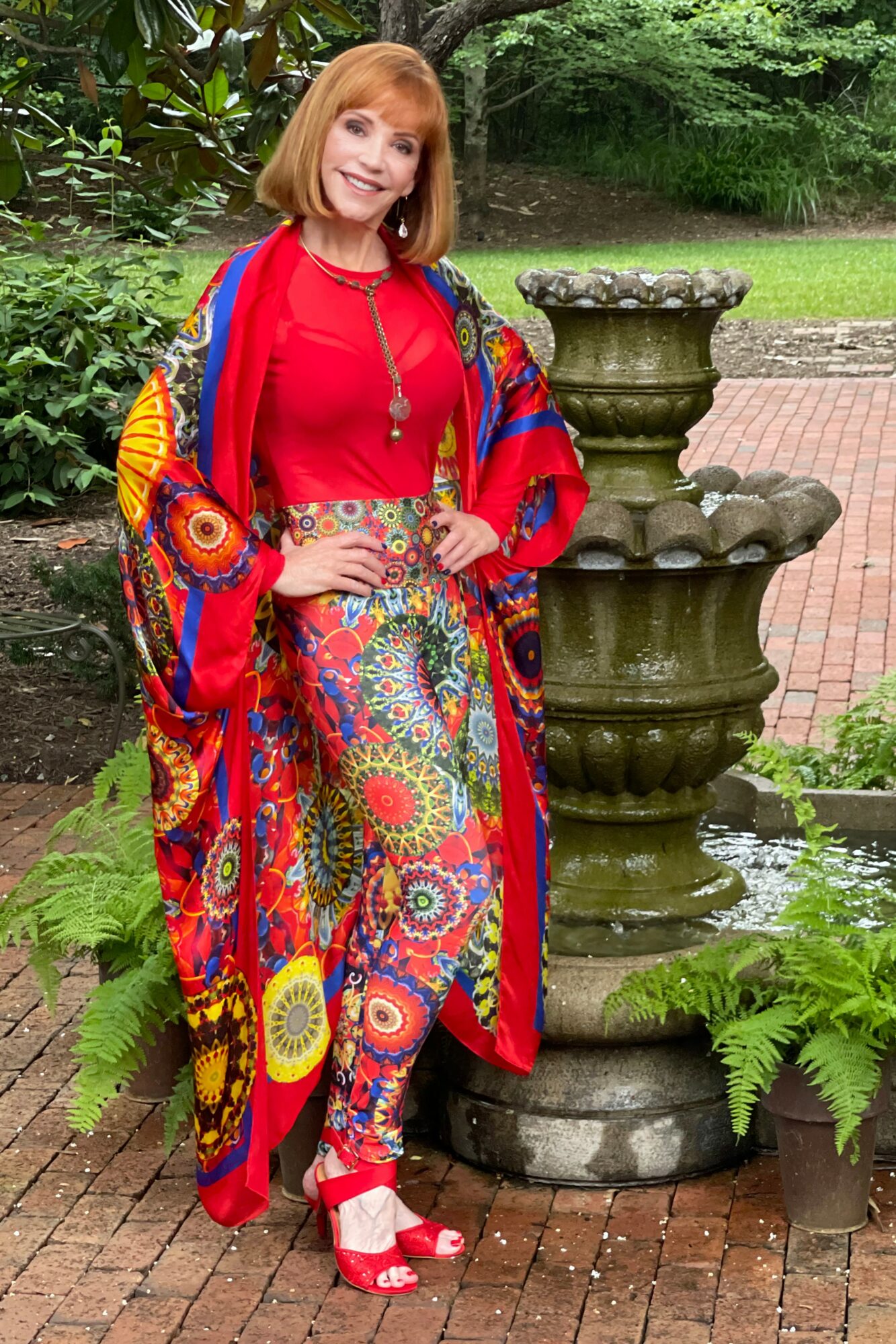 Woman with red hair in a colorful outfit standing next to a fountain in a garden, smiling, with greenery around.