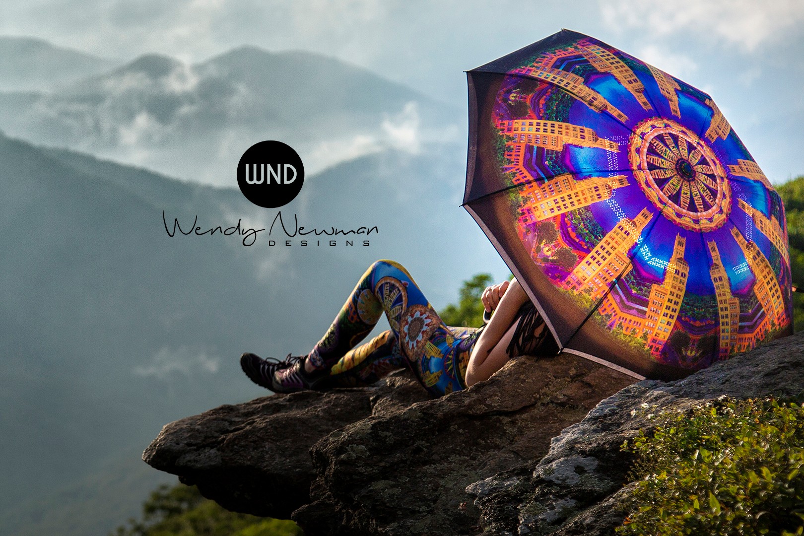 Person lying on a rock holding a colorful umbrella with a mountain landscape background.