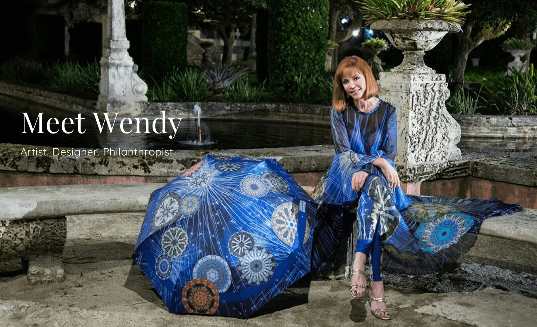 Woman sitting outdoors with a blue umbrella and matching clothing, surrounded by stone structures and greenery.