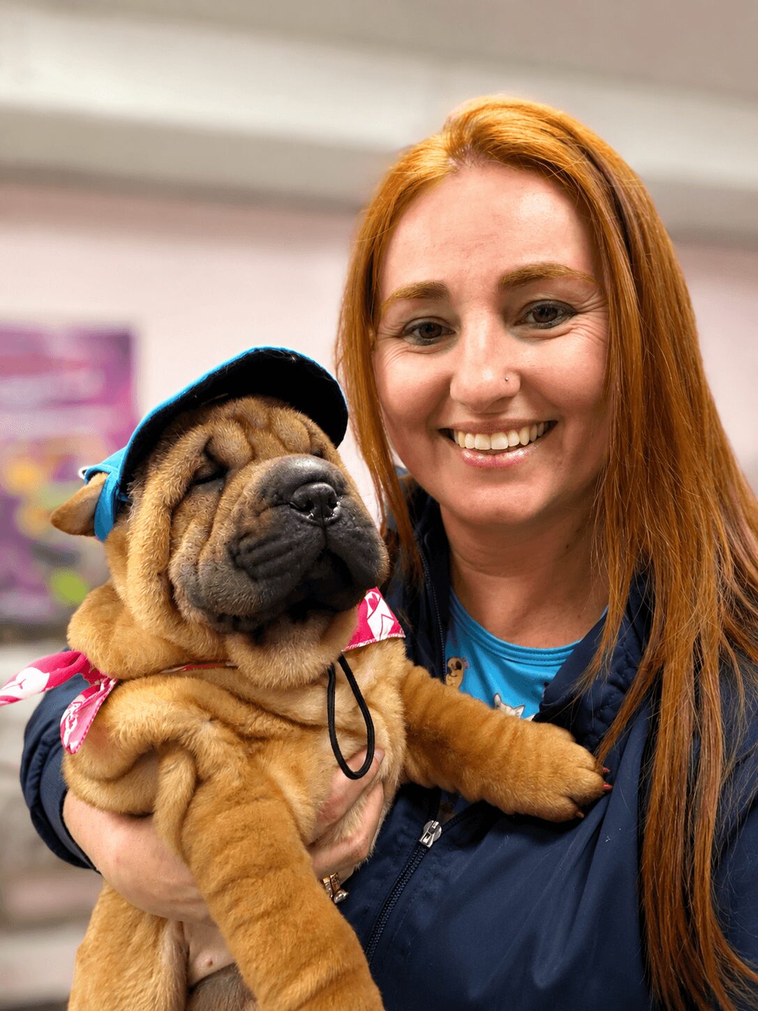 Woman with long red hair smiling while holding a puppy wearing a helmet, indoors.