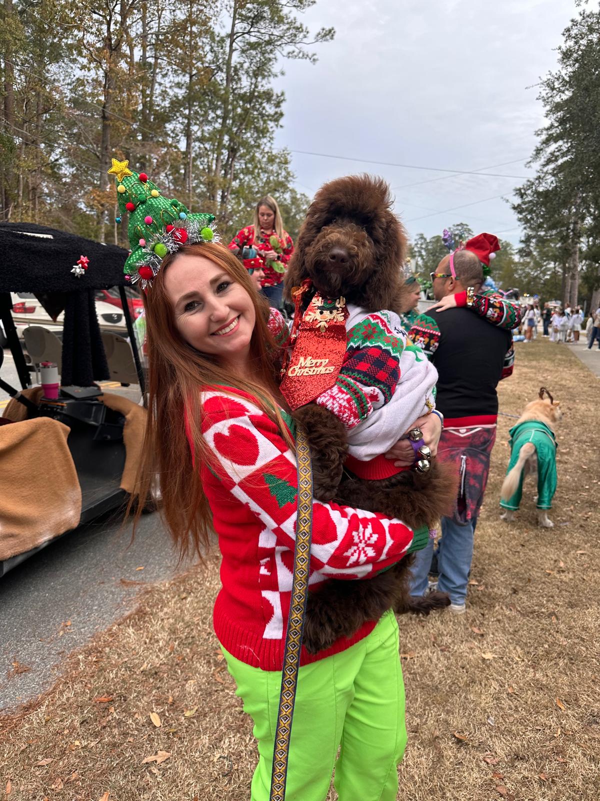 Woman in Christmas sweater holding a large dog dressed in holiday clothing outdoors during a festive event.