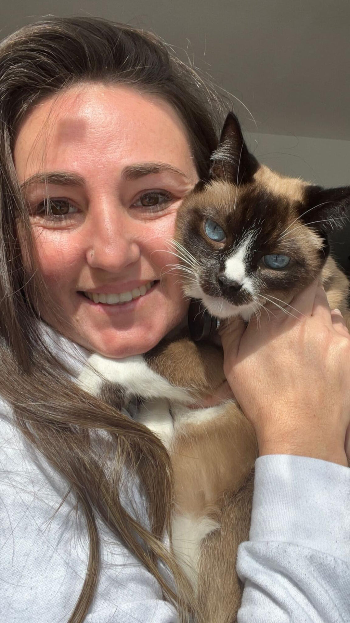 Woman smiling holding a Siamese cat with blue eyes close to her face.