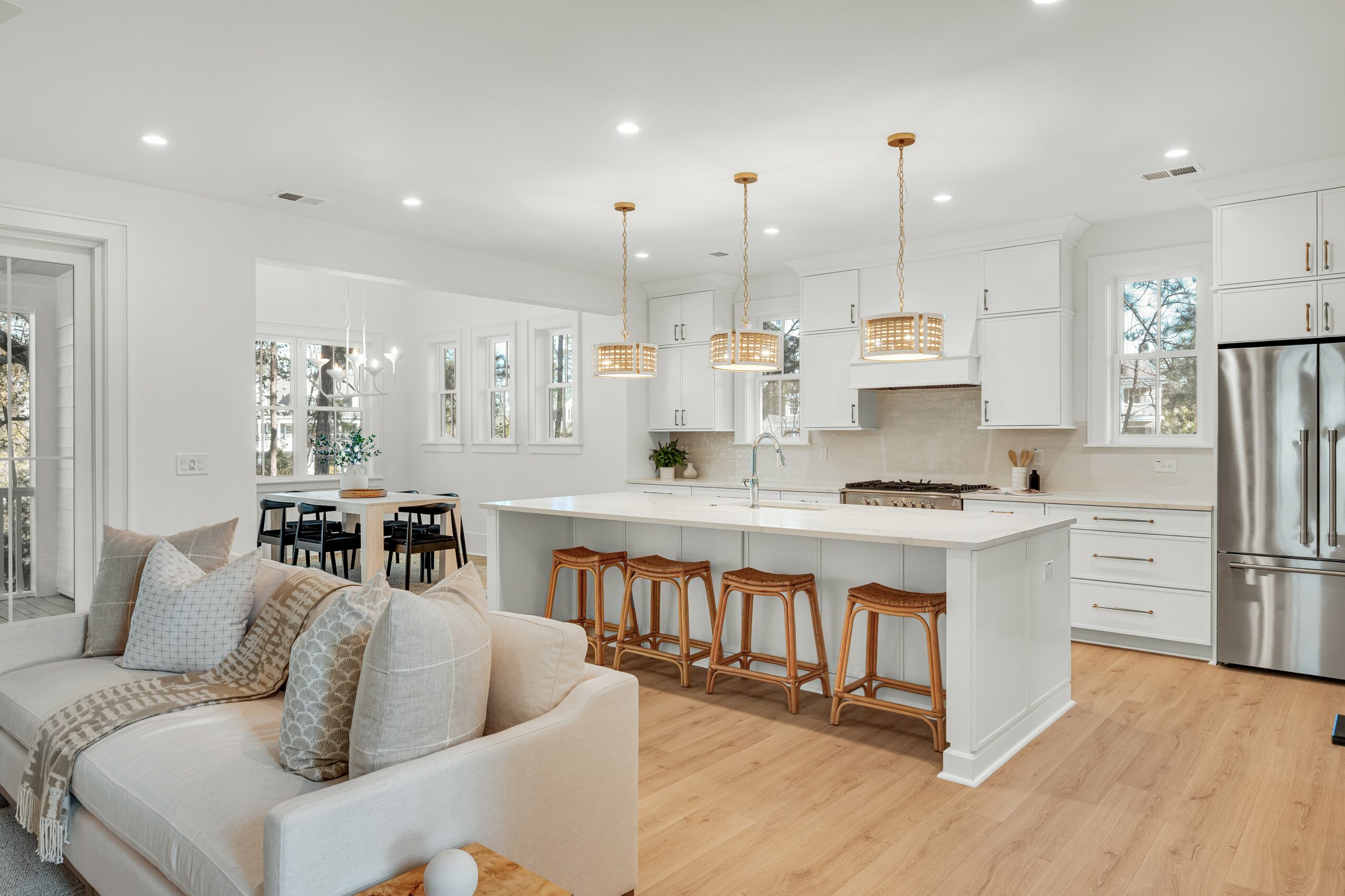 Open kitchen and living room with white cabinets, island, pendant lights, dining table, and sofa, hardwood floors, large windows, and stainless steel refrigerator.