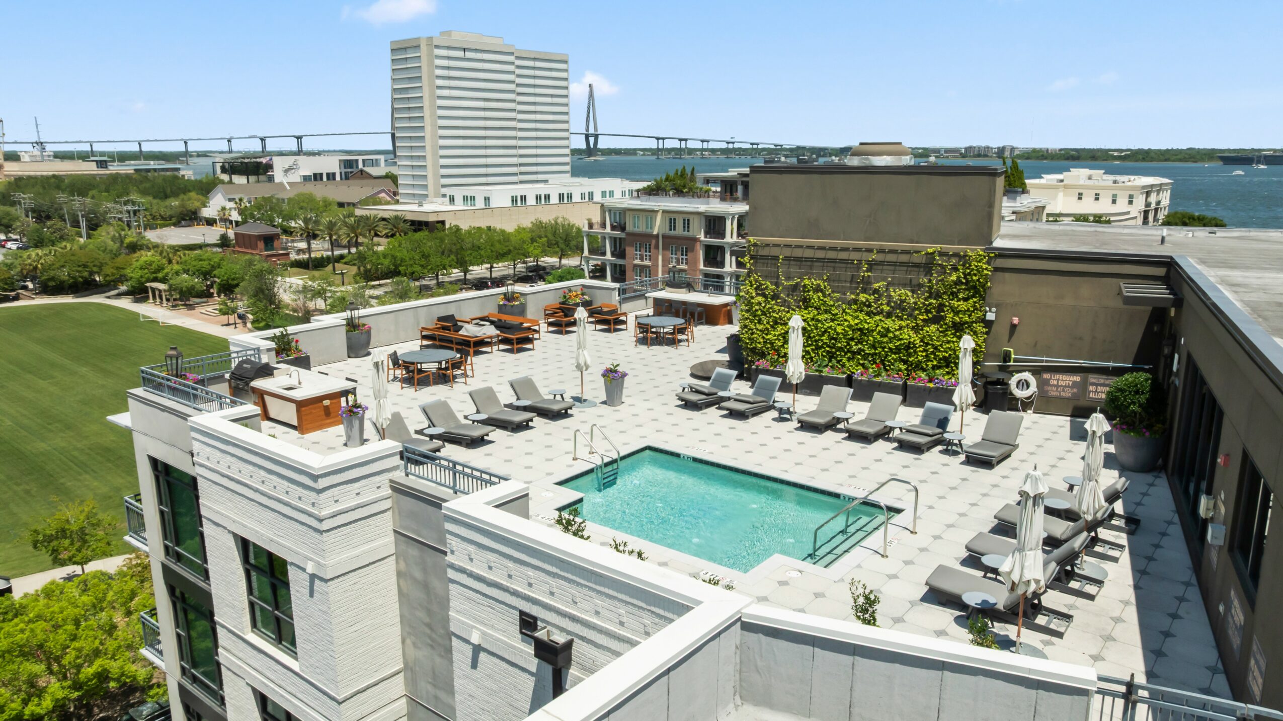 Rooftop pool area with lounge chairs, umbrellas, and surrounding buildings, overlooking a body of water and cityscape.