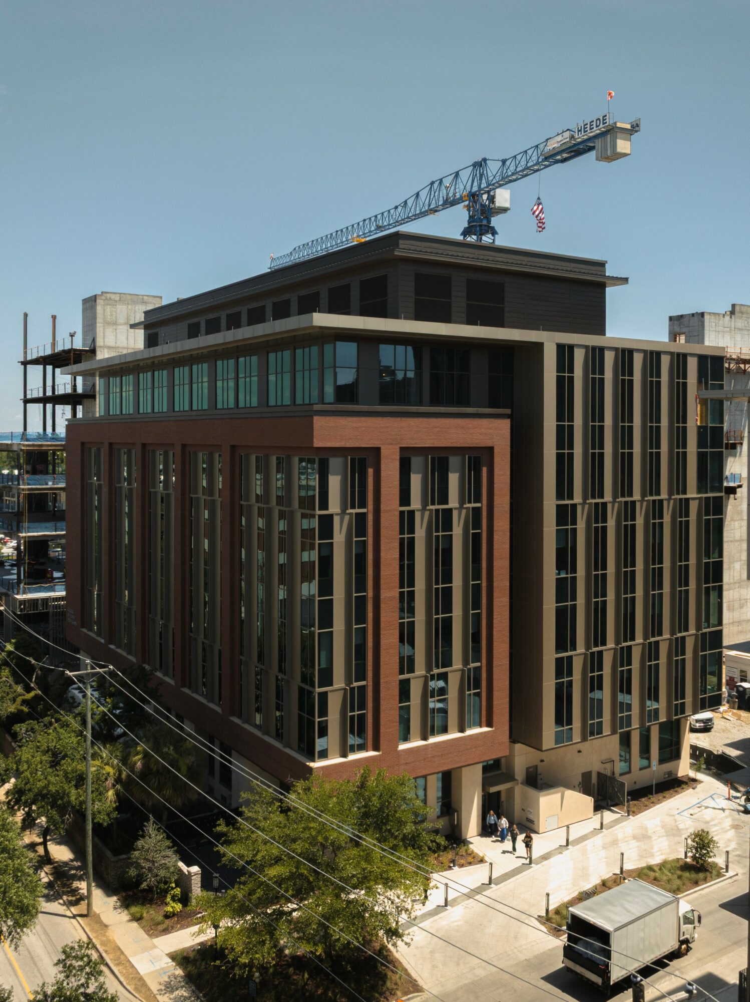 Modern multi-story building with glass windows and a construction crane above, situated on a city street with trees and parked vehicles.
