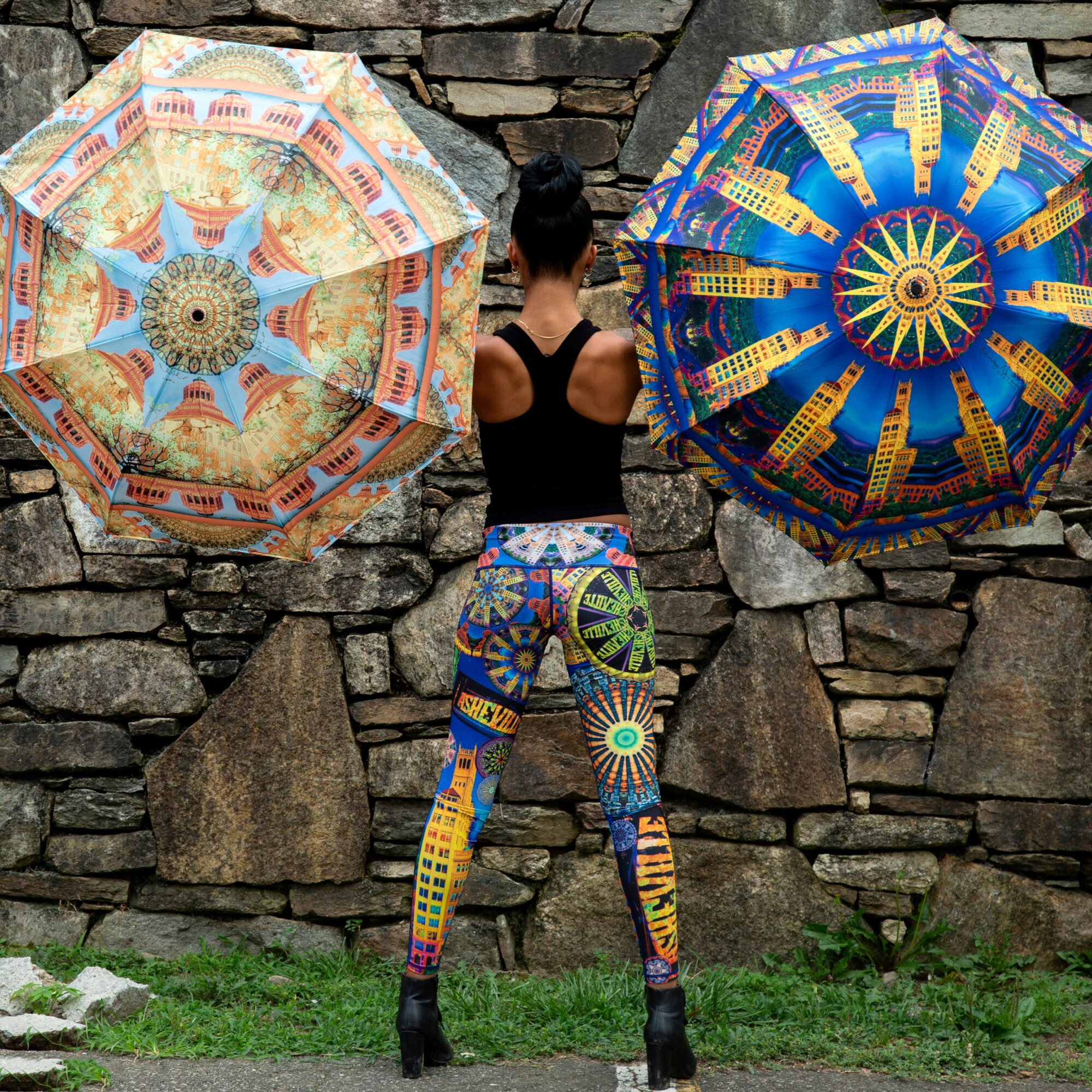 Person standing between two colorful umbrellas against a stone wall, wearing patterned leggings and a black top.