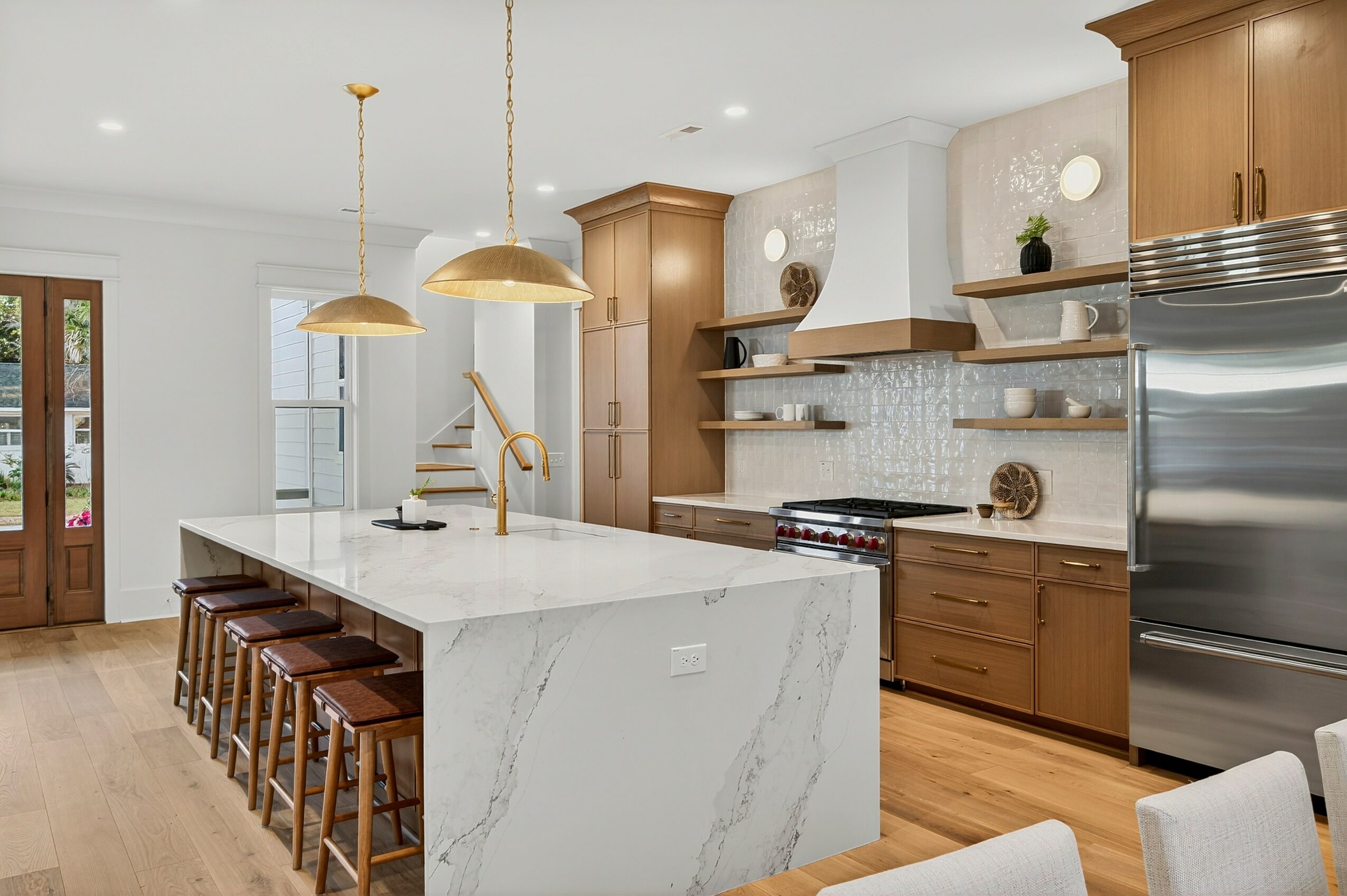 Modern kitchen with white island, wooden cabinets, stainless steel appliances, and pendant lights, with a staircase in the background.