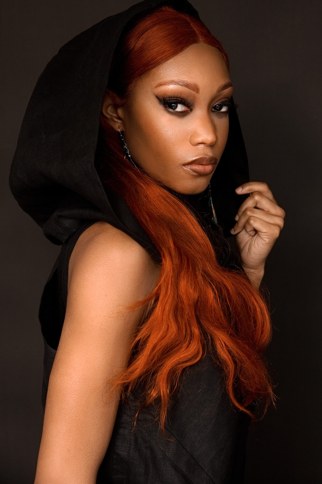 Woman with long red hair and dark makeup, wearing a black hooded garment, looking over shoulder against dark background.